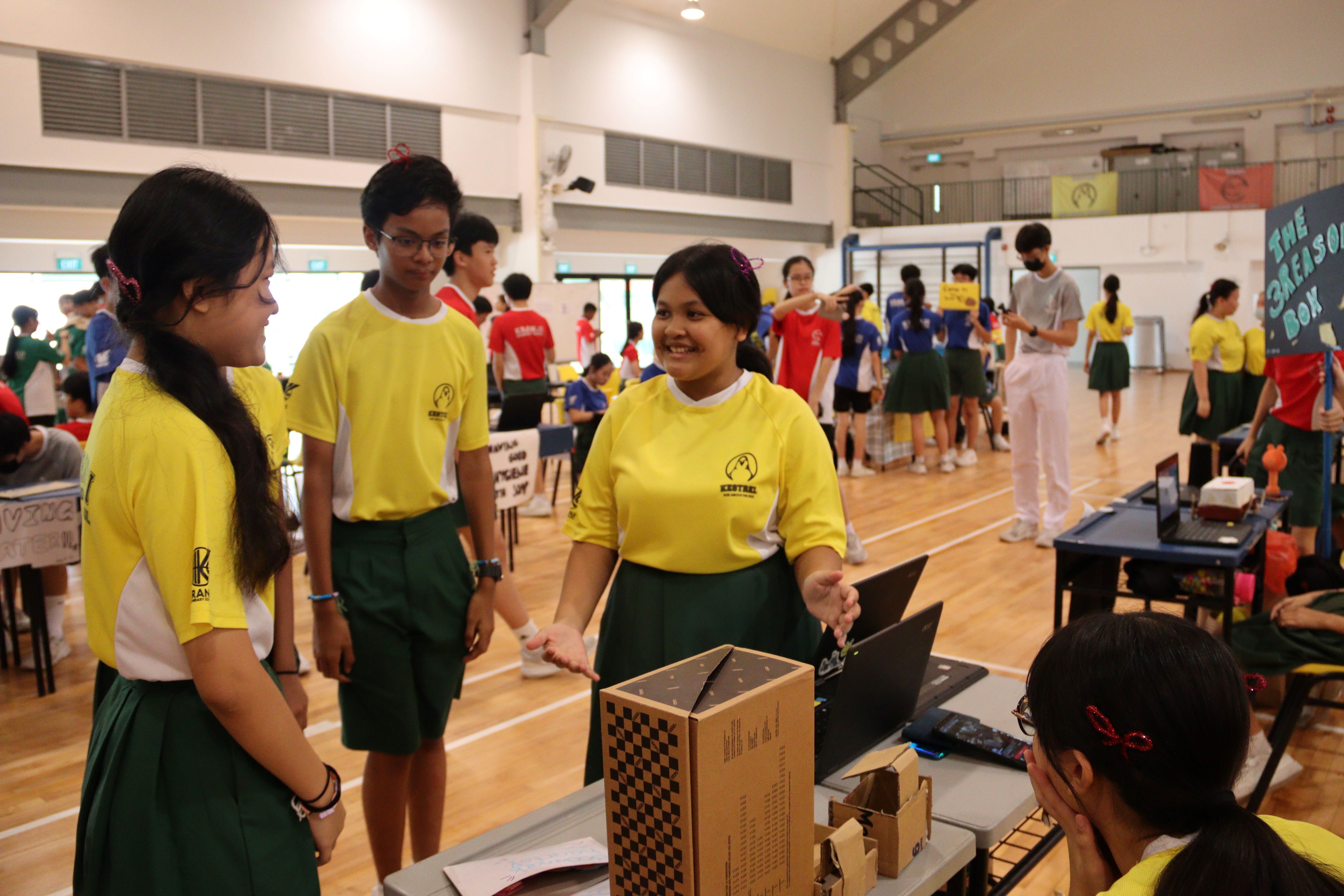 Students in yellow and green uniforms present a cardboard project at a school exhibition in a large hall.