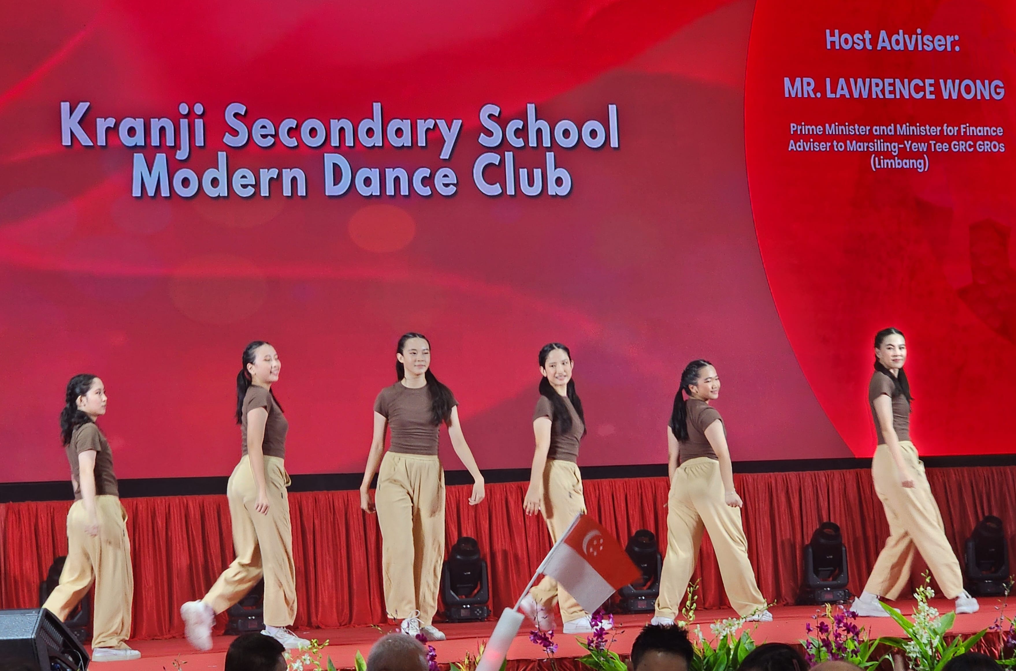 Six students in brown tops and tan pants perform on stage for Kranji Secondary School Modern Dance Club.