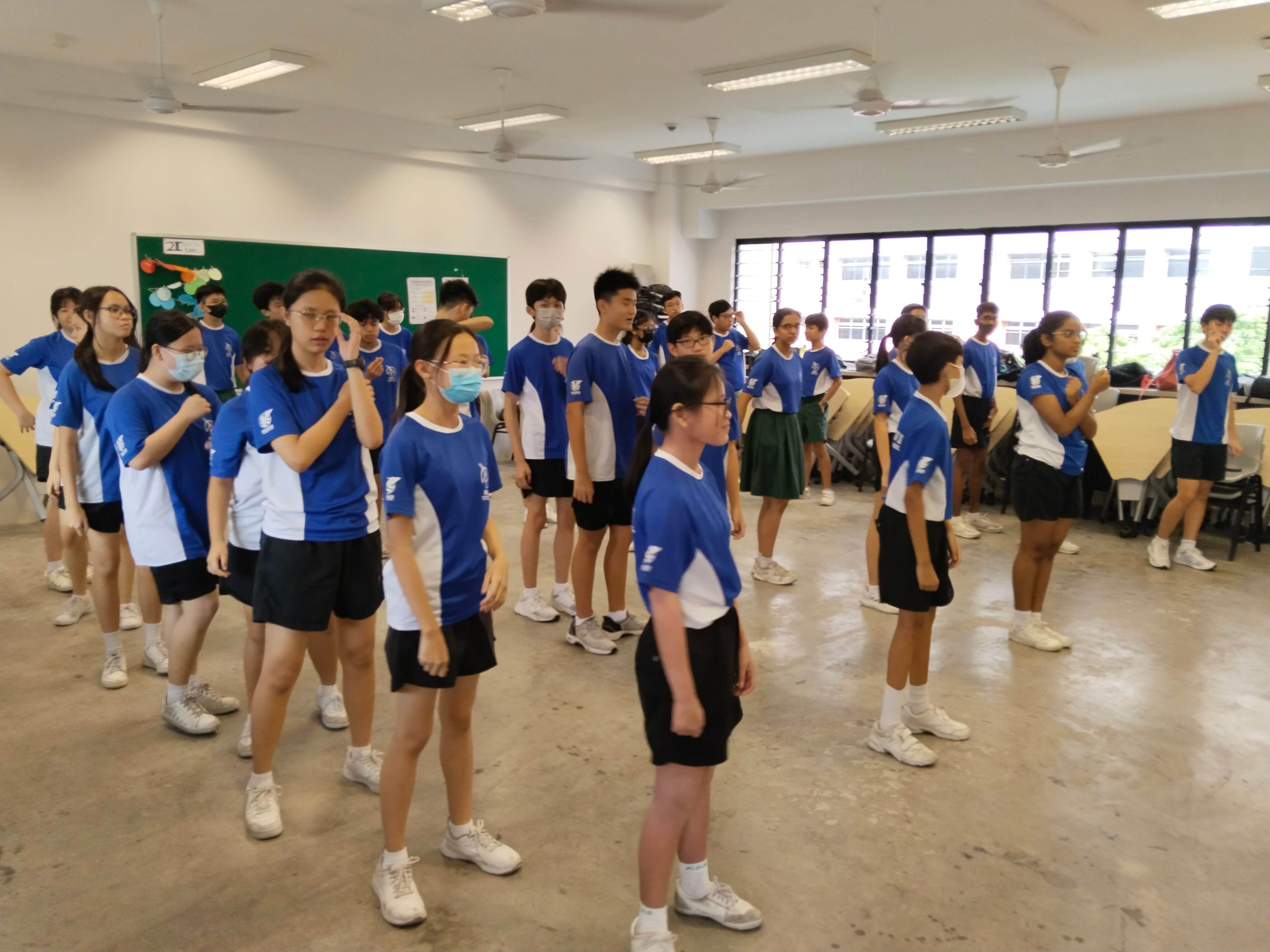 A large group of students in blue and white uniforms stand in rows inside a school hall, doing an activity.