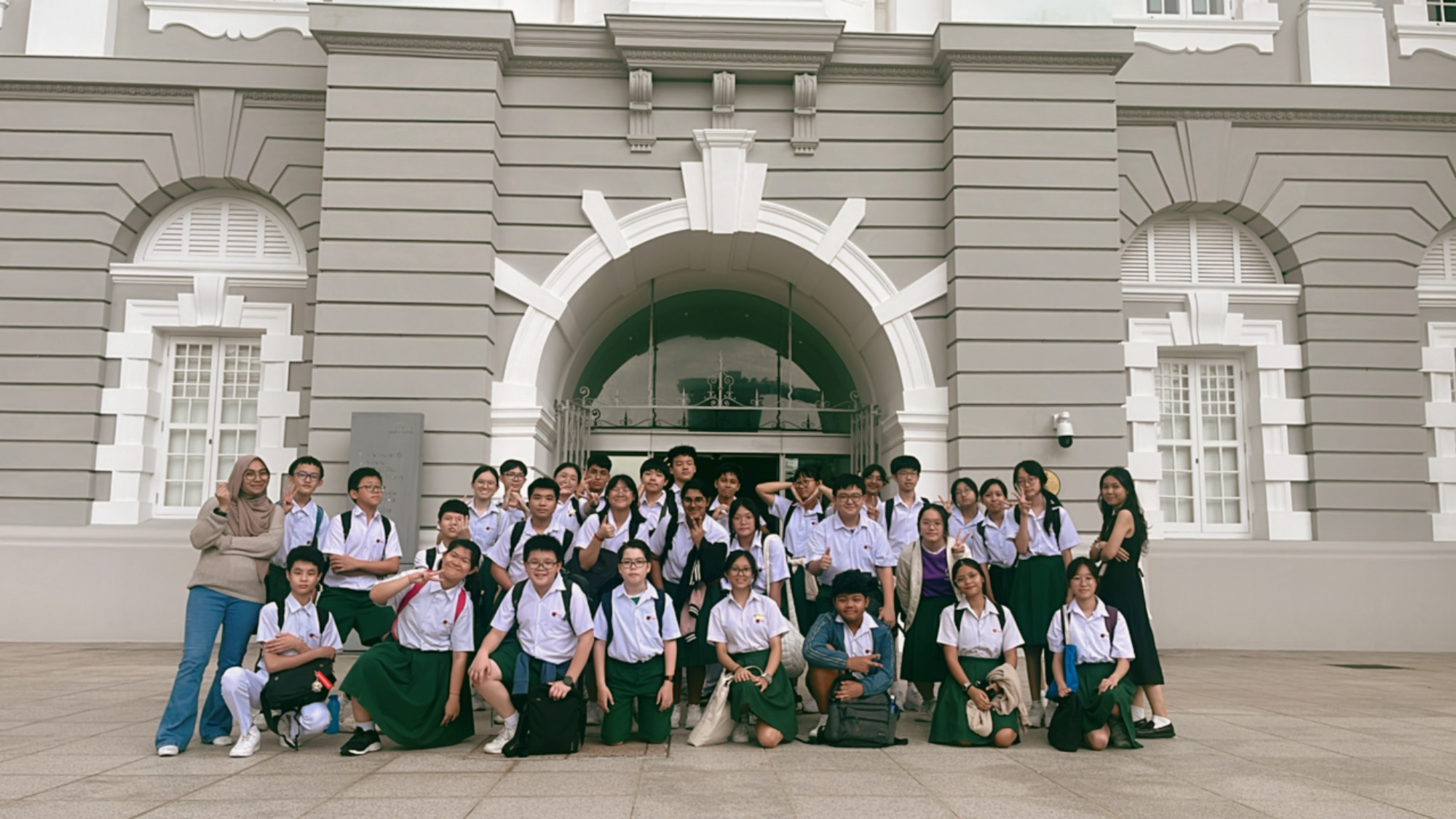 A large group of students in white shirts and green skirts pose in front of a grand grey colonial building with an arched entrance.