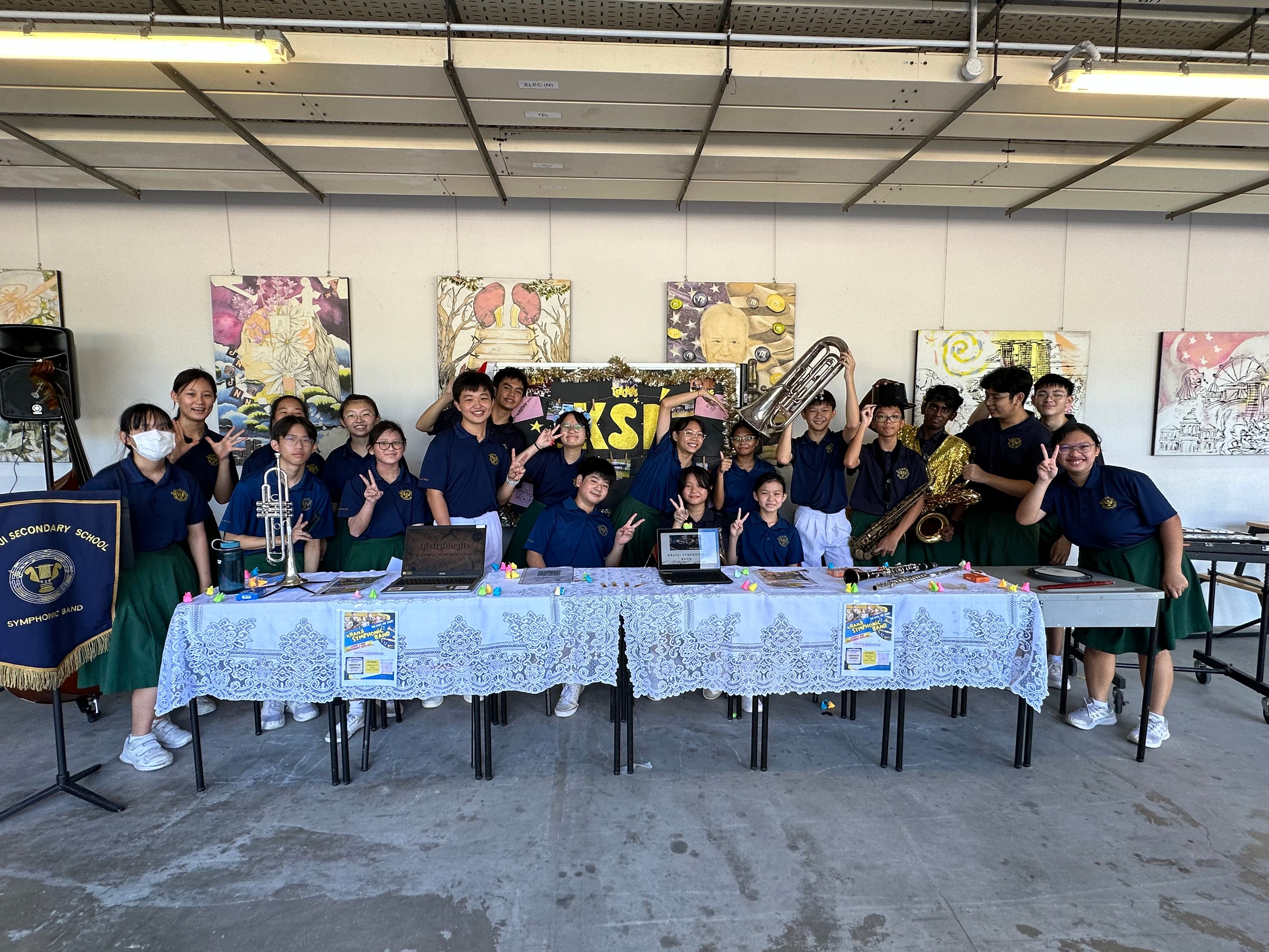 Students in navy uniforms pose with brass instruments at a lace-covered table under a Symphonic Band banner.