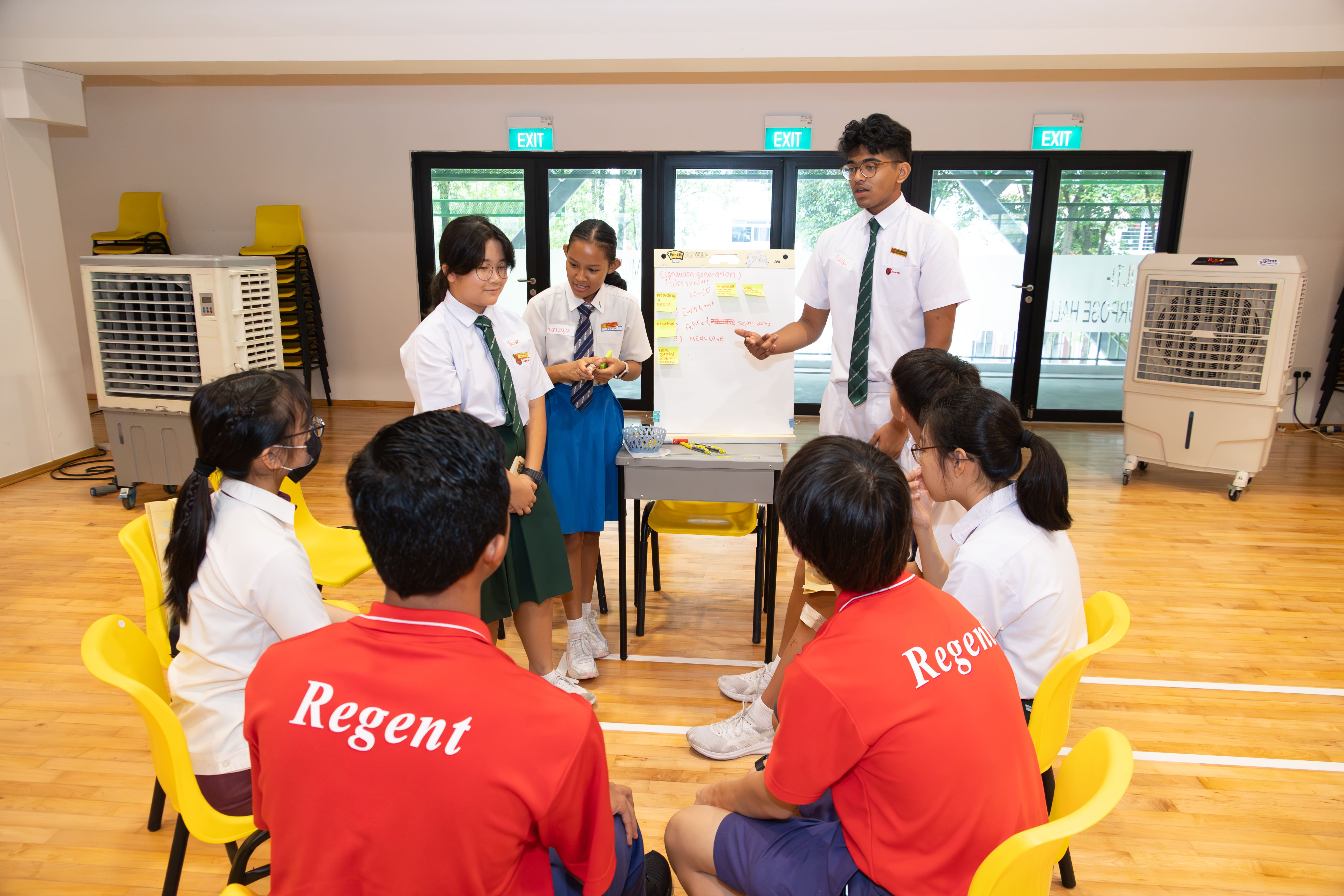 Students in school uniforms sit in a circle while two peers present at a whiteboard with sticky notes in a hall.