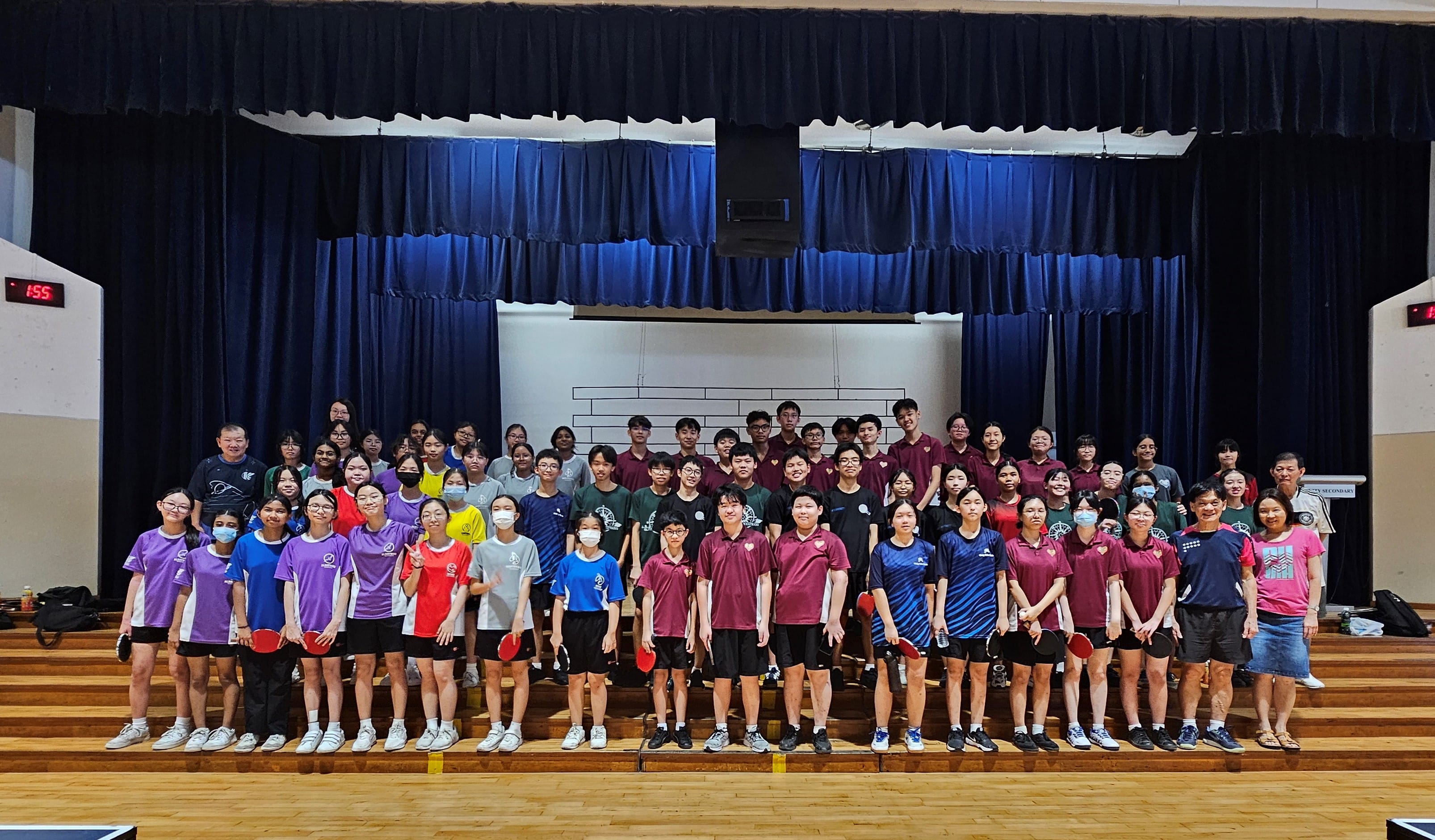 Large group of students in colorful sports jerseys holding table tennis paddles posing together in a school hall.