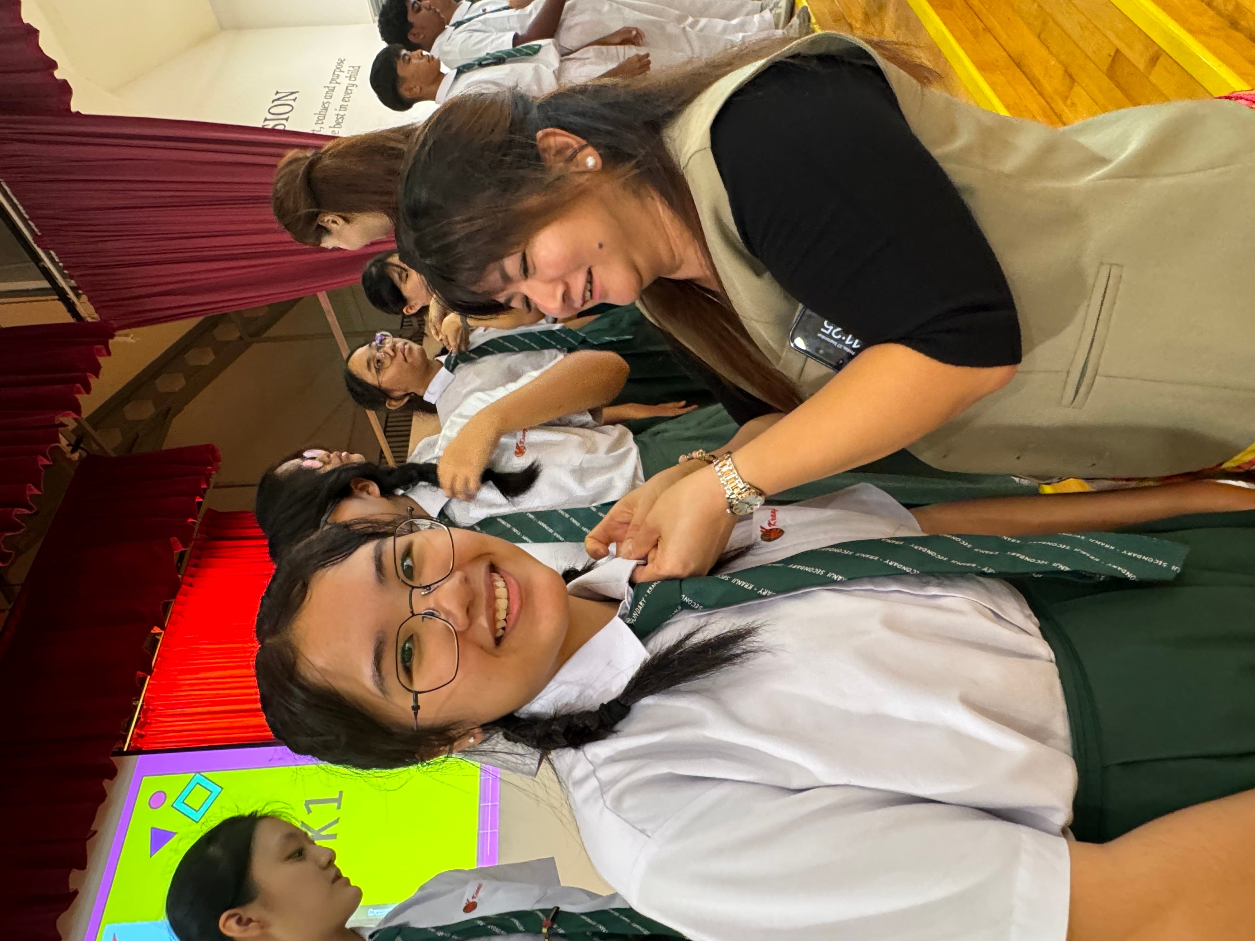 An adult pins a badge on a smiling female student in school uniform with green tie in a hall.