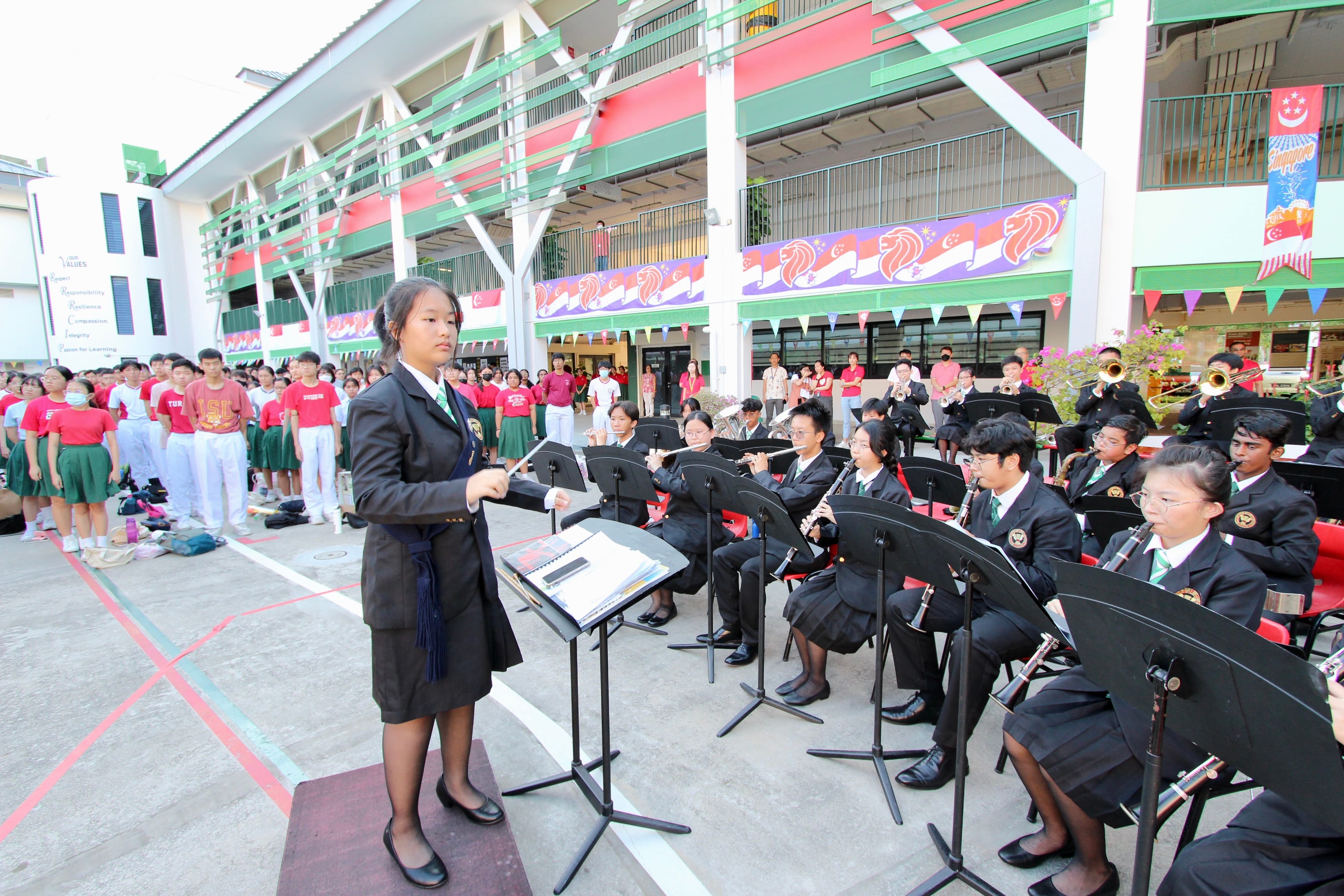 A student in a black uniform conducts a school band outdoors while other students watch in a decorated school courtyard.