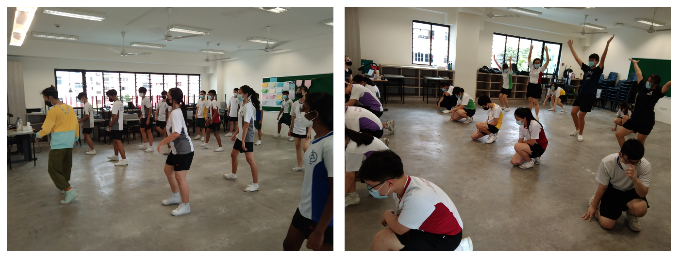 Students wearing masks practice dance or movement exercises in a large indoor room, some standing and some crouching.