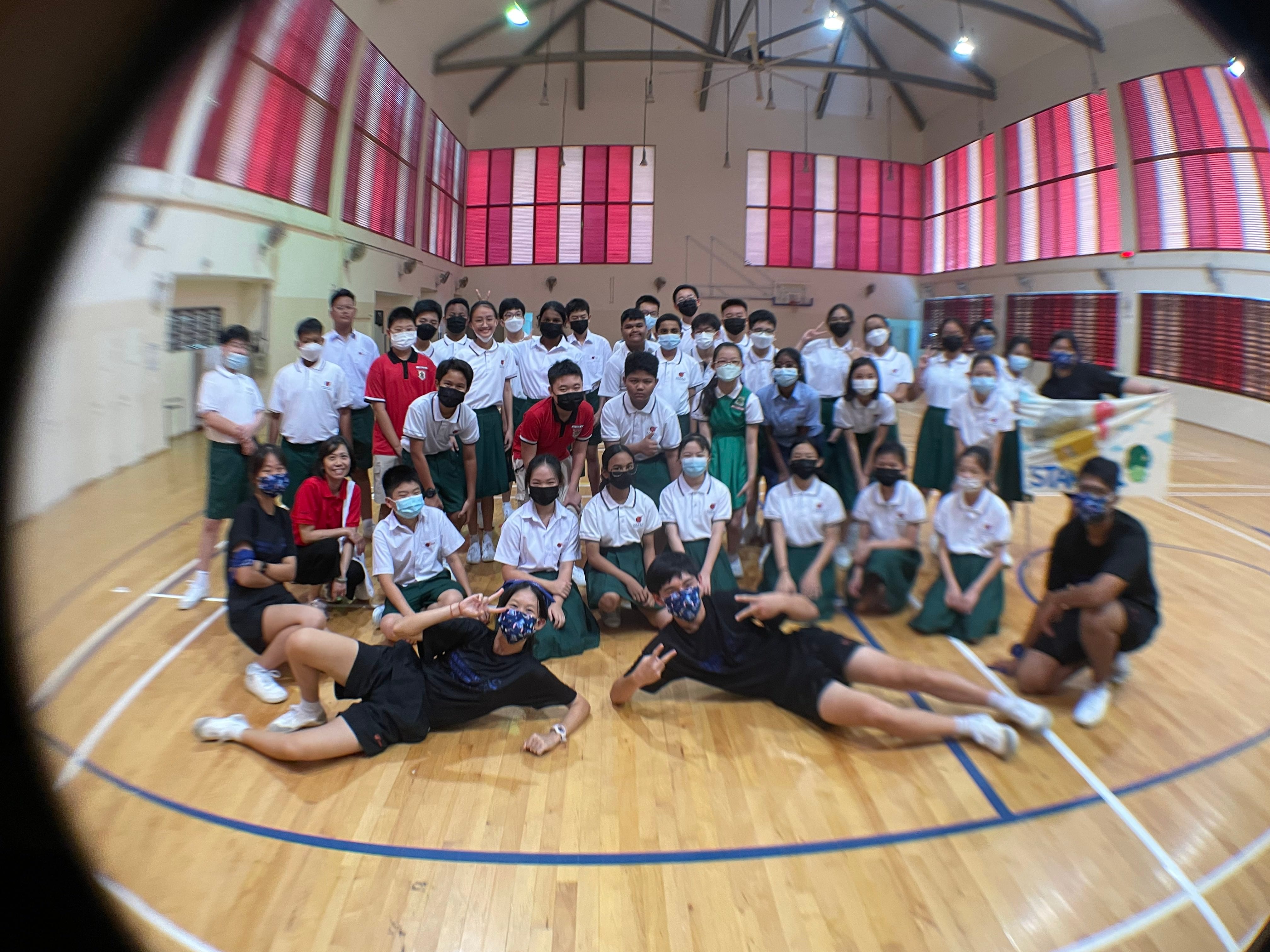 Large group of masked students in school uniforms posing together inside a gymnasium with red-paneled windows.