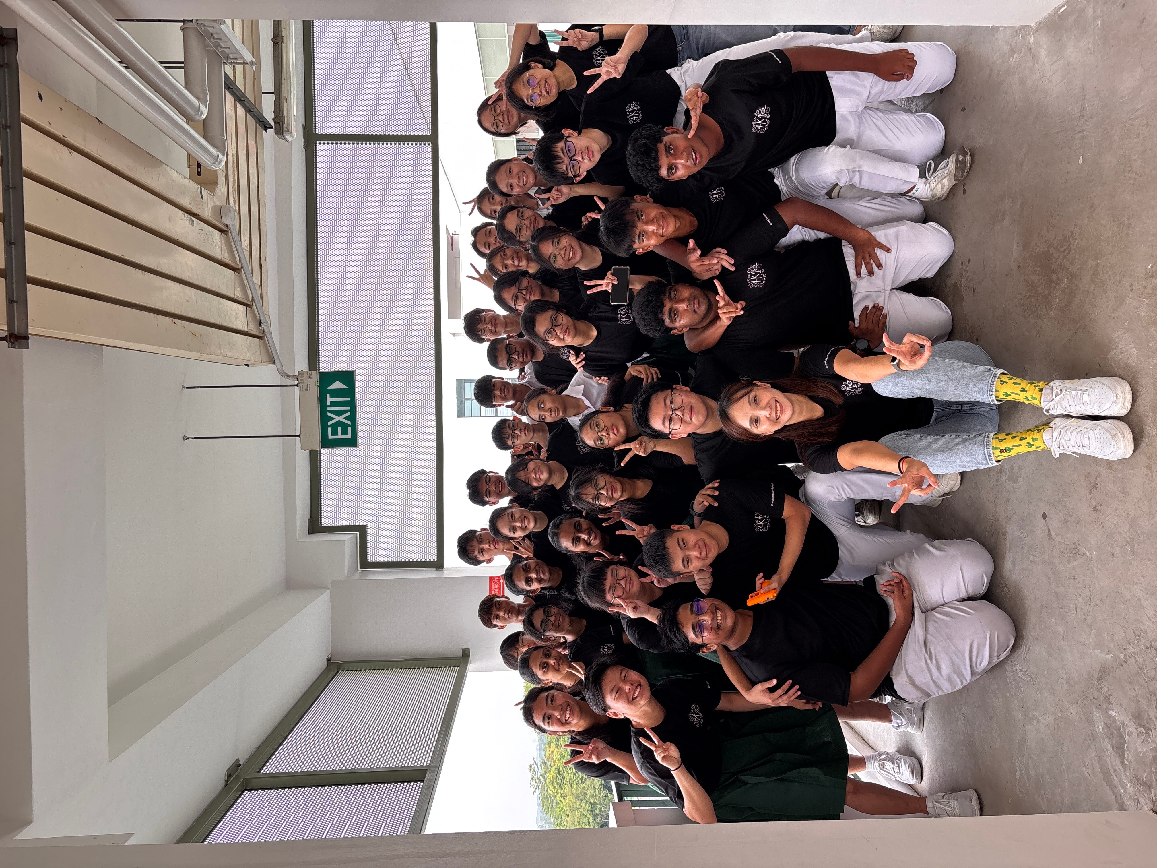Large group in black shirts and white pants pose in a hallway under an EXIT sign, flashing peace signs.