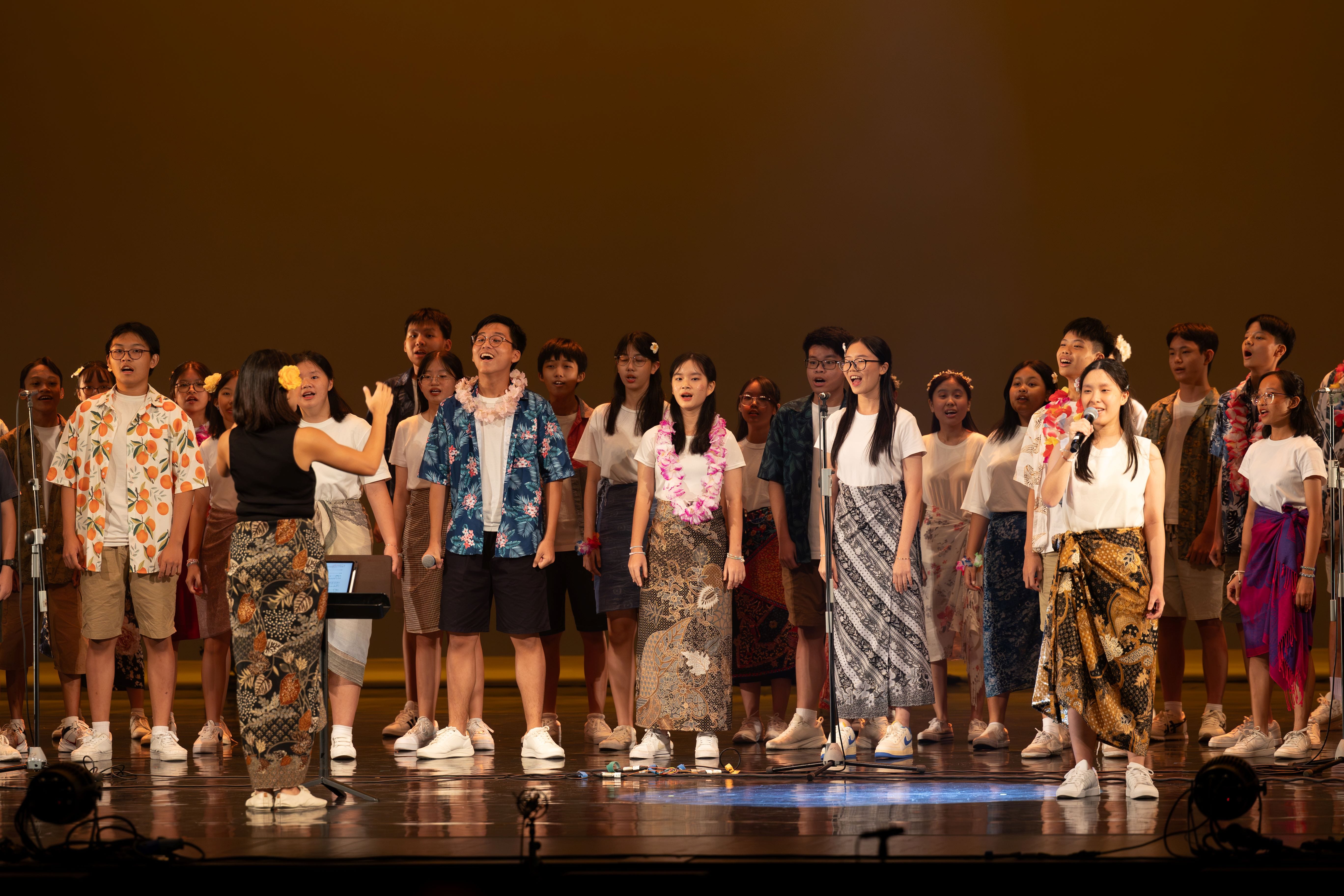 A large youth choir in tropical and batik outfits performs on stage, with a conductor and solo singers at microphones.