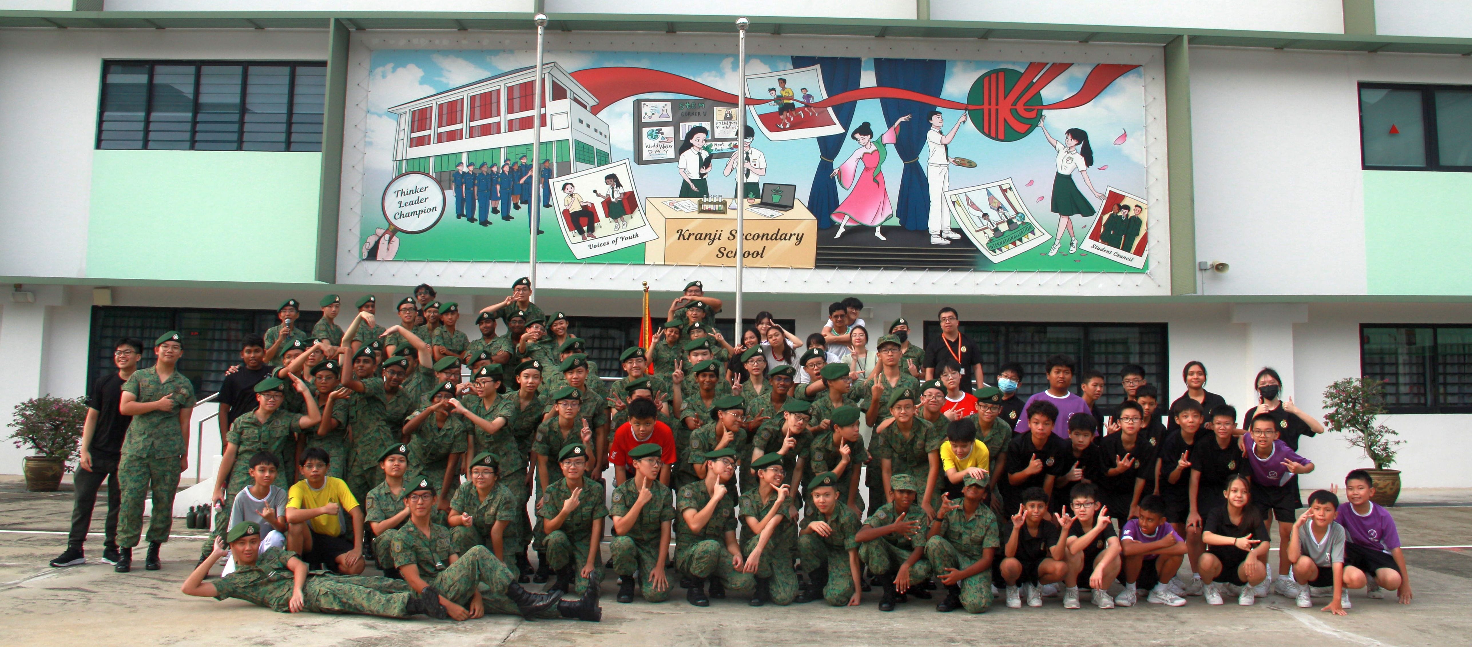 Large group of students in camouflage uniforms posing in front of Kranji Secondary School's colorful mural.