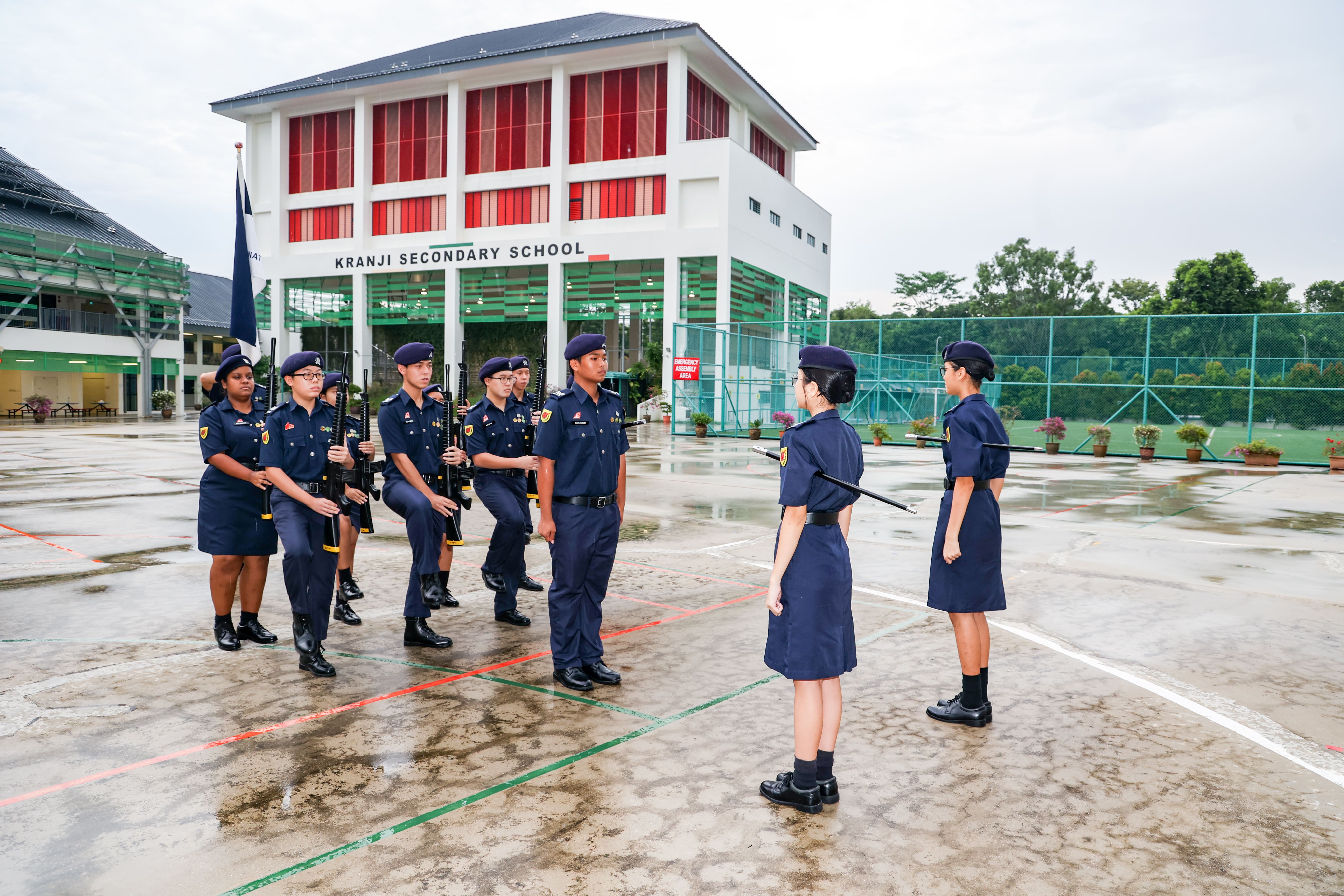 Students in navy uniforms with rifles drill on a wet courtyard in front of Kranji Secondary School building.