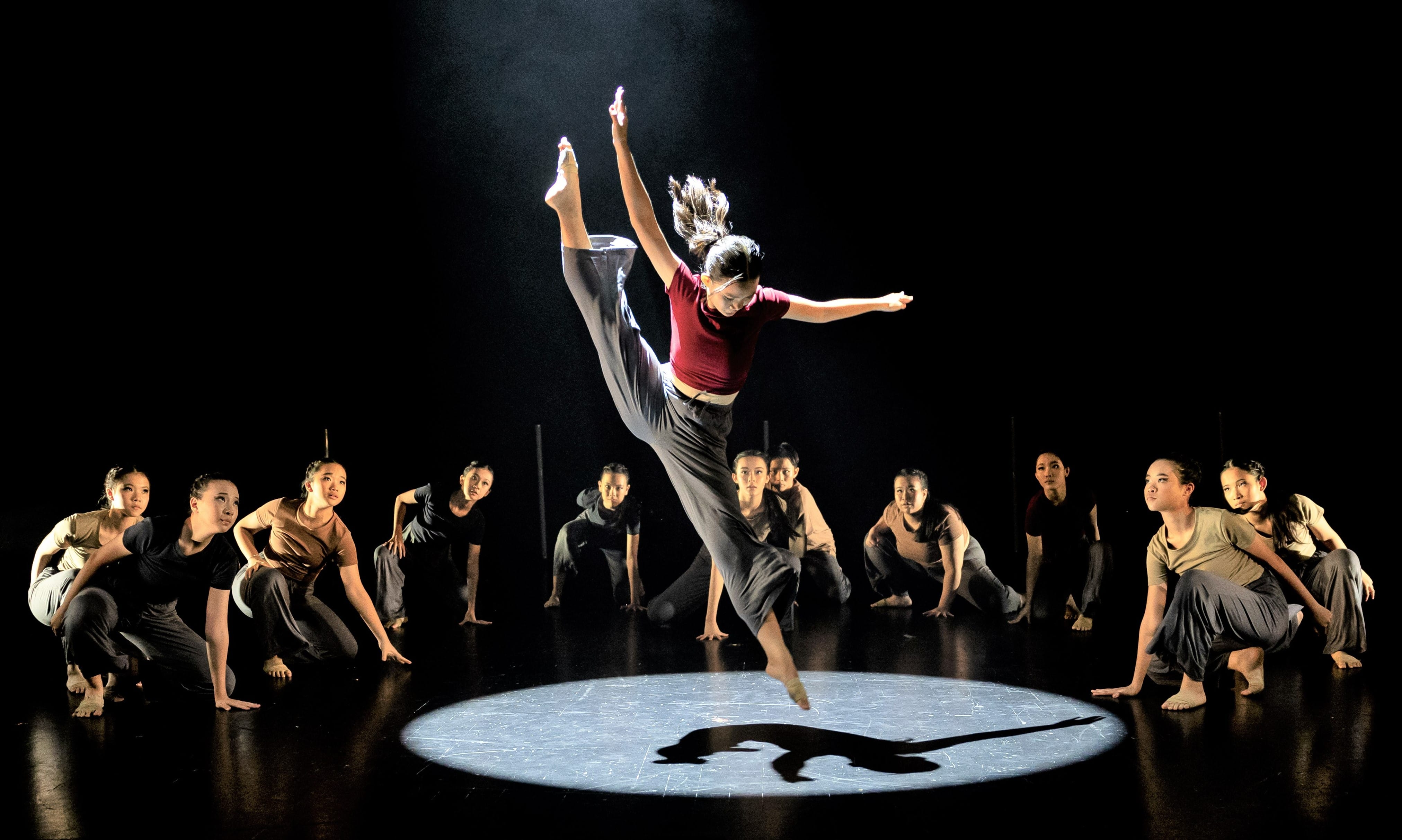 A dancer leaps in a spotlight on a dark stage while a group of dancers crouch in a circle around her.