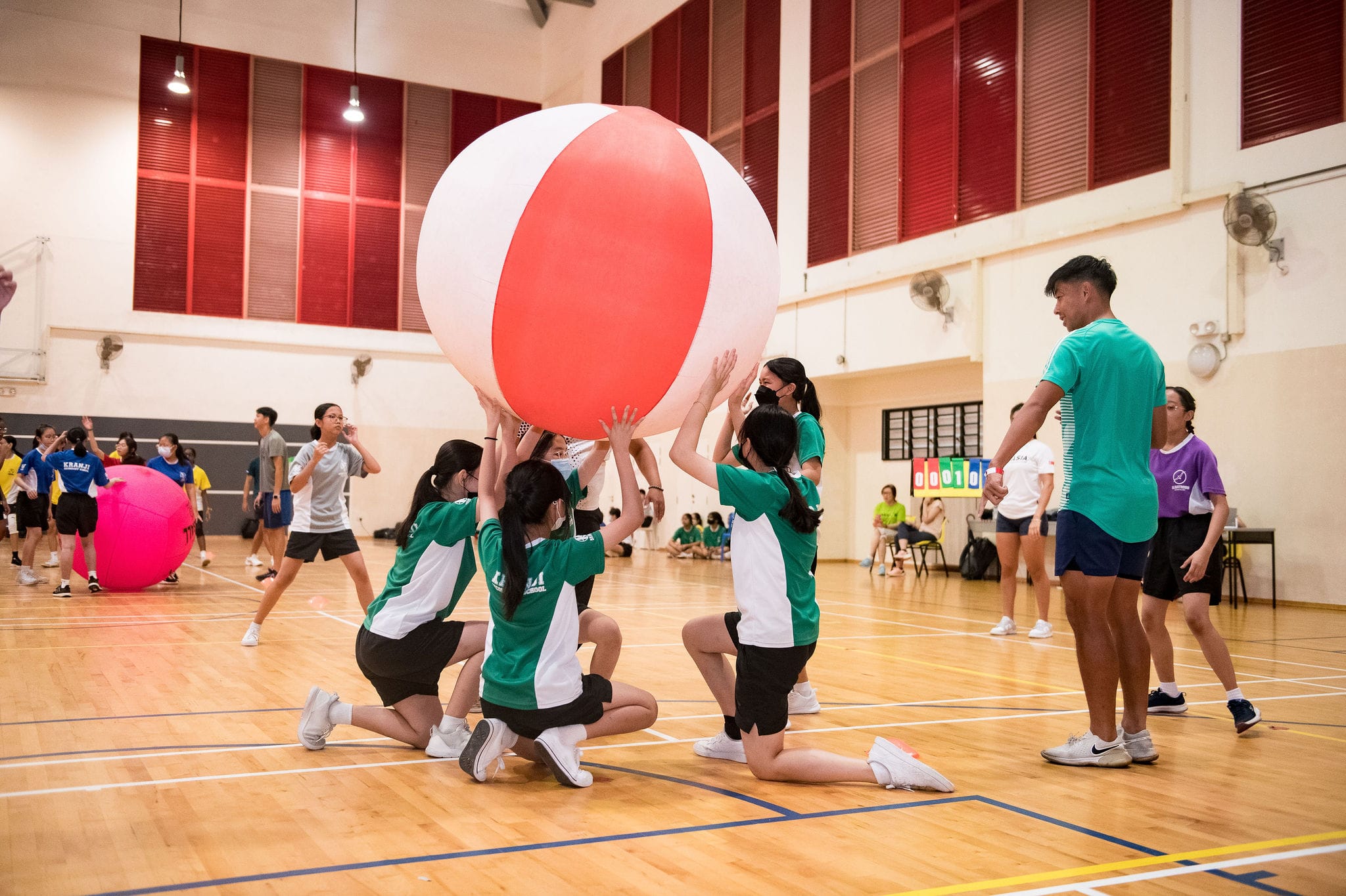 Students in green shirts lift a large red and white inflatable ball together on an indoor gymnasium court.