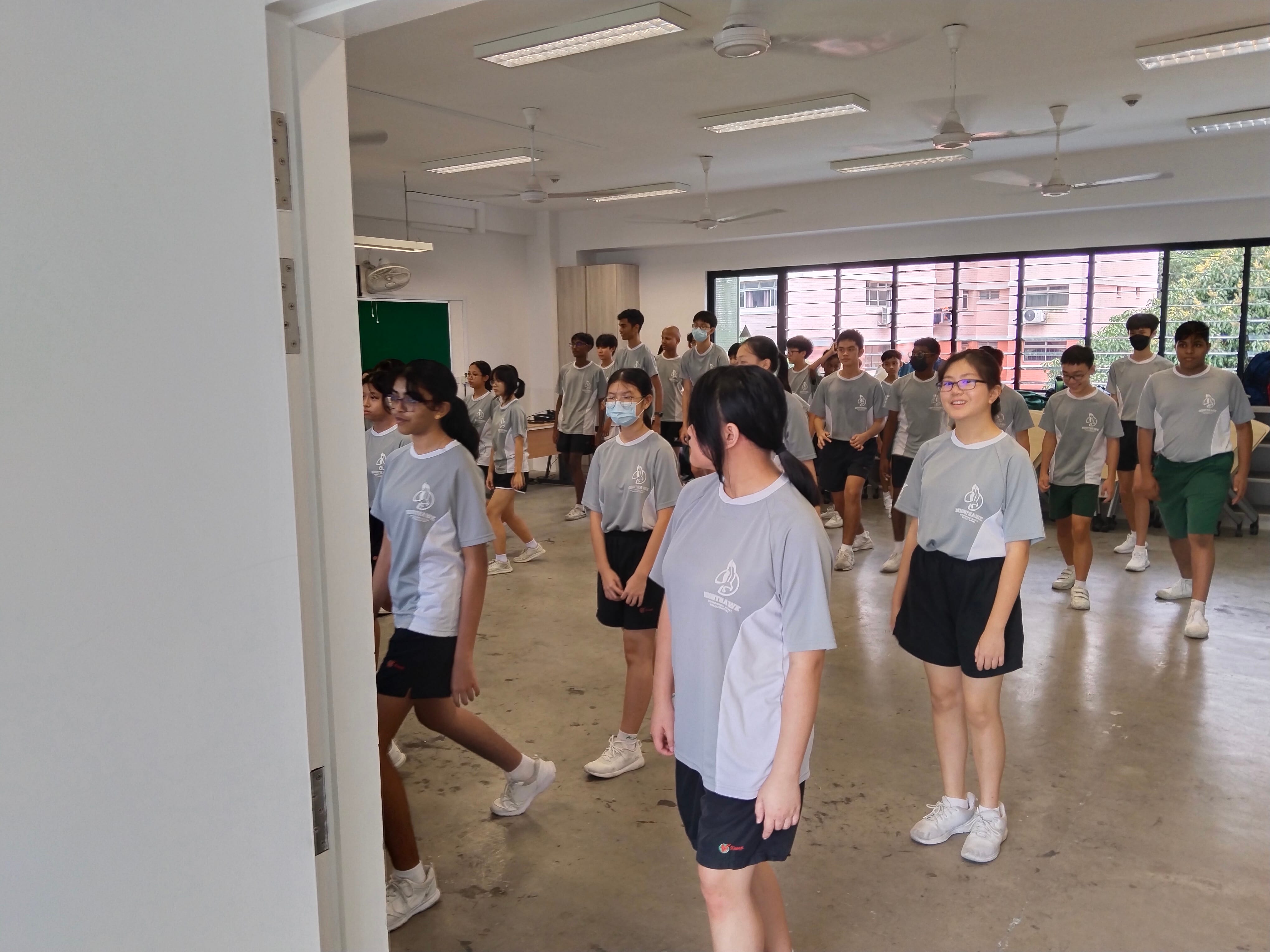 Students in matching gray uniforms stand and move around a large indoor room with ceiling fans and fluorescent lights.