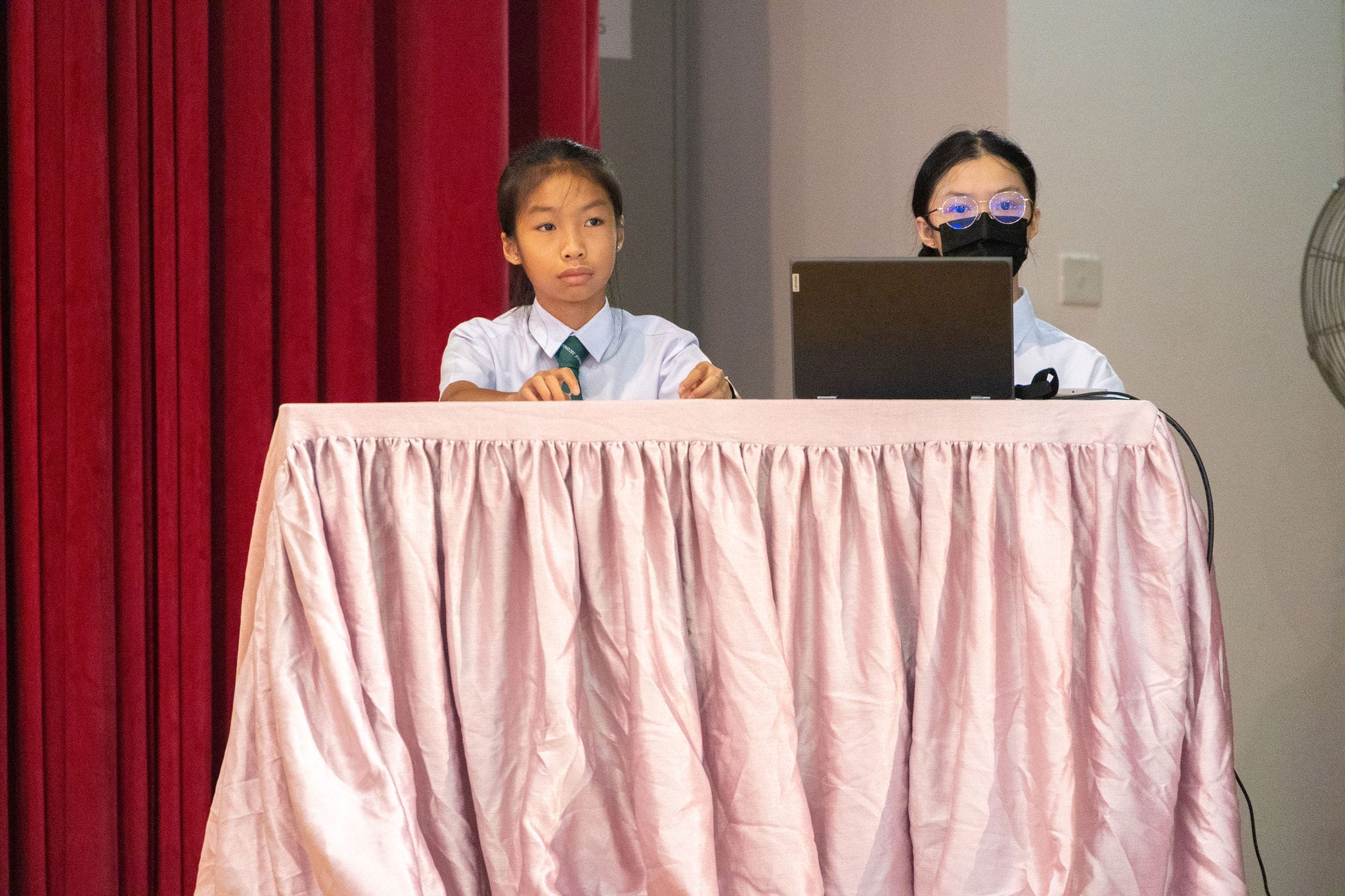 Two uniformed schoolgirls stand behind a pink skirted table, one holding a pen and one with a laptop and face mask.