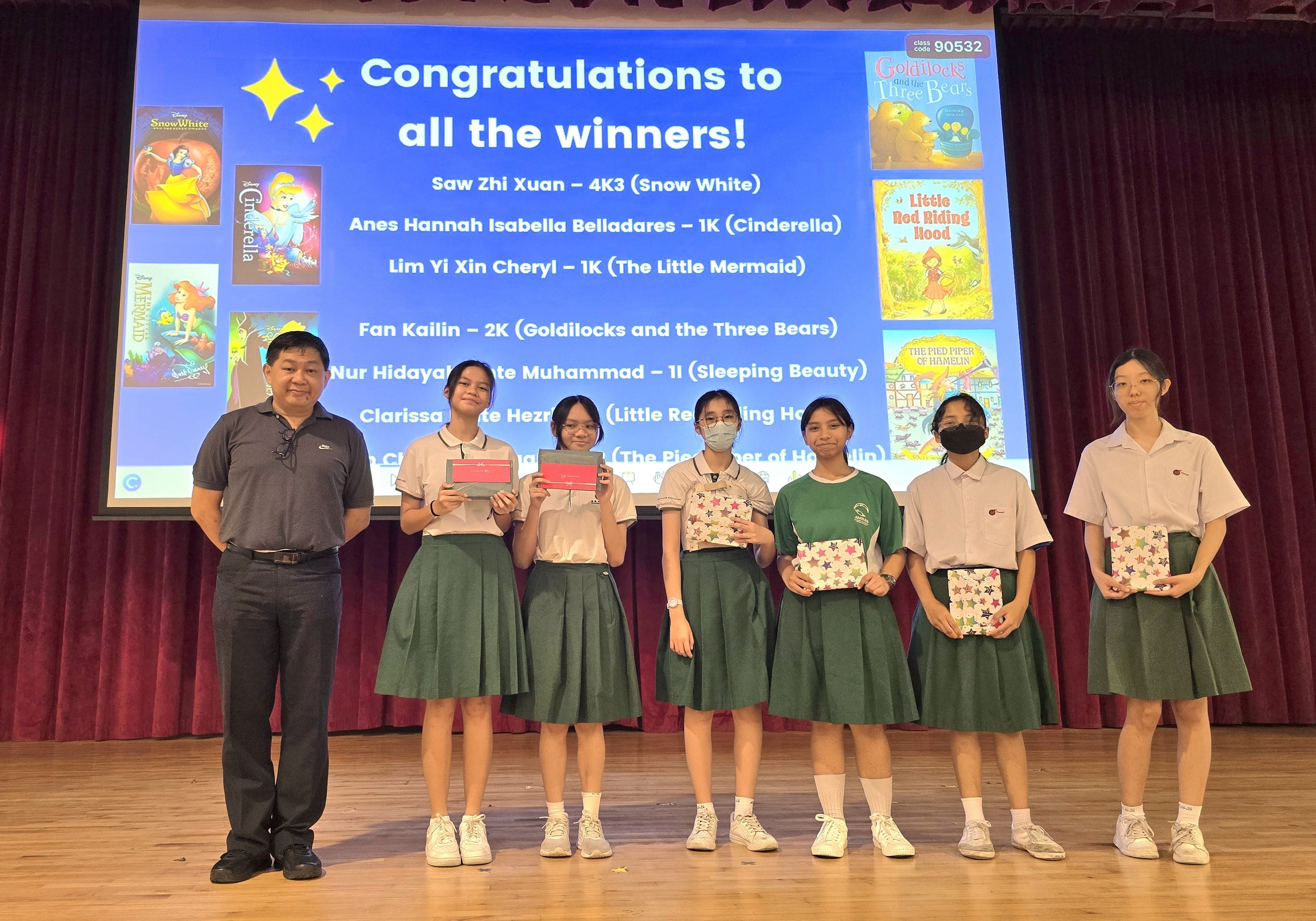 A teacher and five female students in green skirts hold prizes on stage before a congratulations winners slideshow.