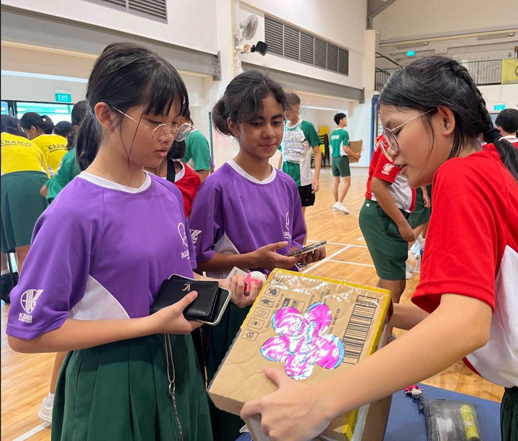 Three school girls in purple and red shirts examine a decorated cardboard box in a school hall.
