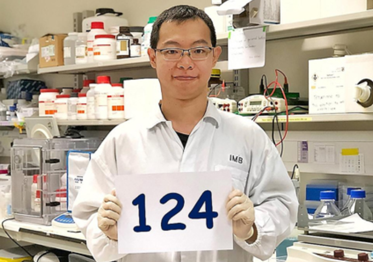 A scientist in a white lab coat holds a sign with the number 124 in a laboratory setting.