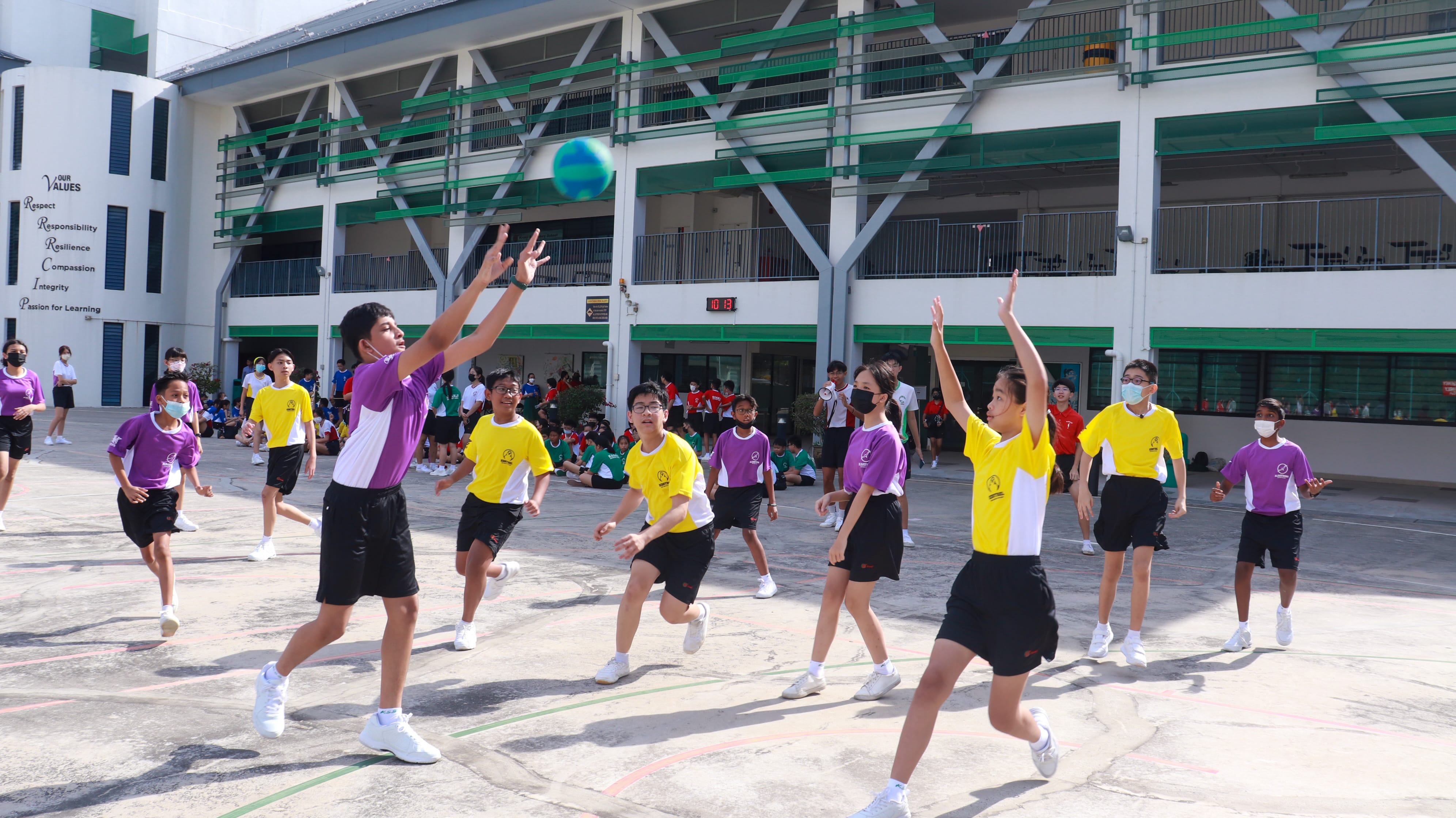 Students in yellow and purple uniforms play handball on a school courtyard with spectators watching nearby.