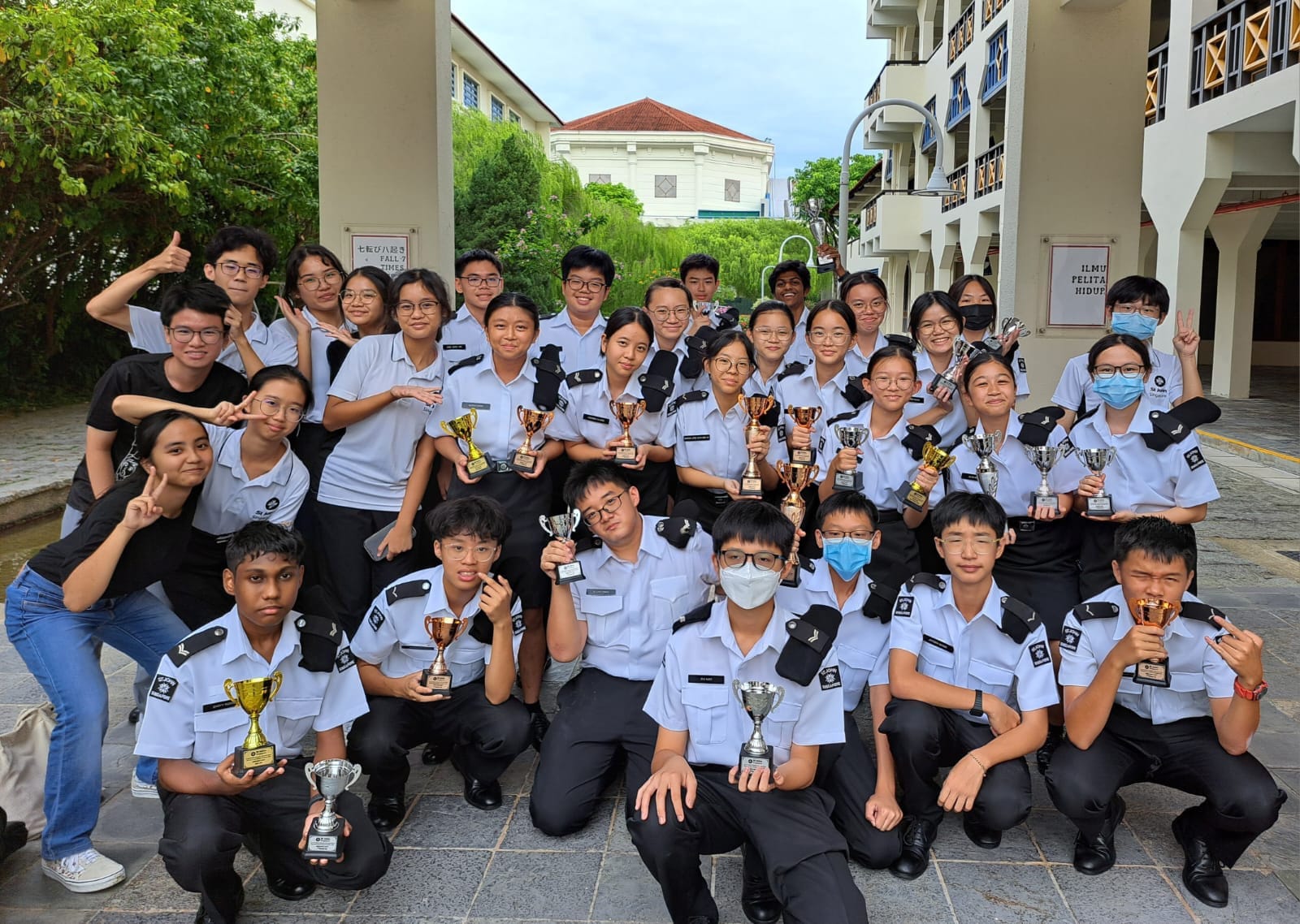 A large group of students in white and black uniforms posing outdoors, holding multiple gold and silver trophies.