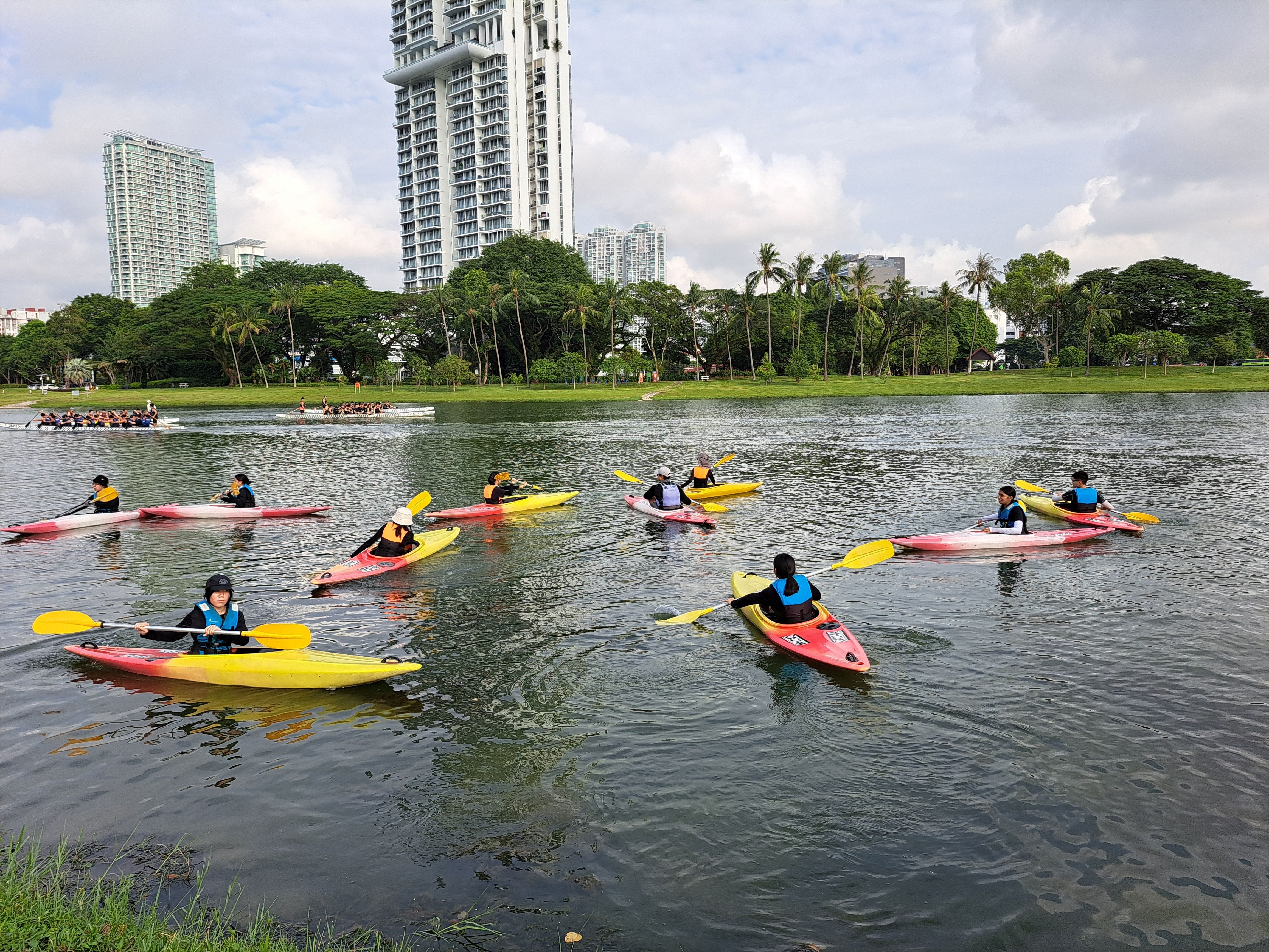 Several people in life vests paddling yellow and red kayaks on a calm urban lake with tall buildings behind.