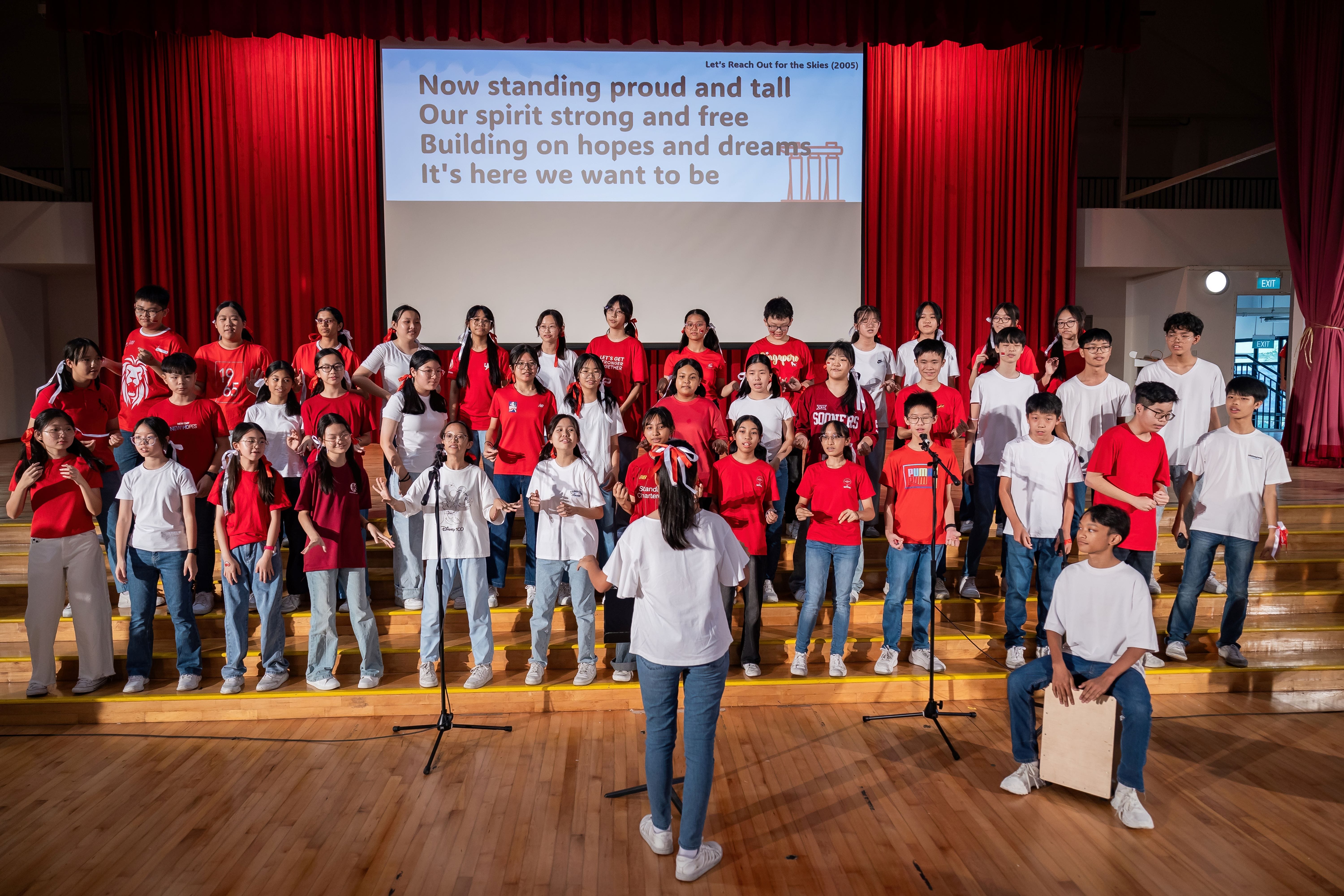 A student choir in red and white shirts performs on a school stage with song lyrics projected behind them.