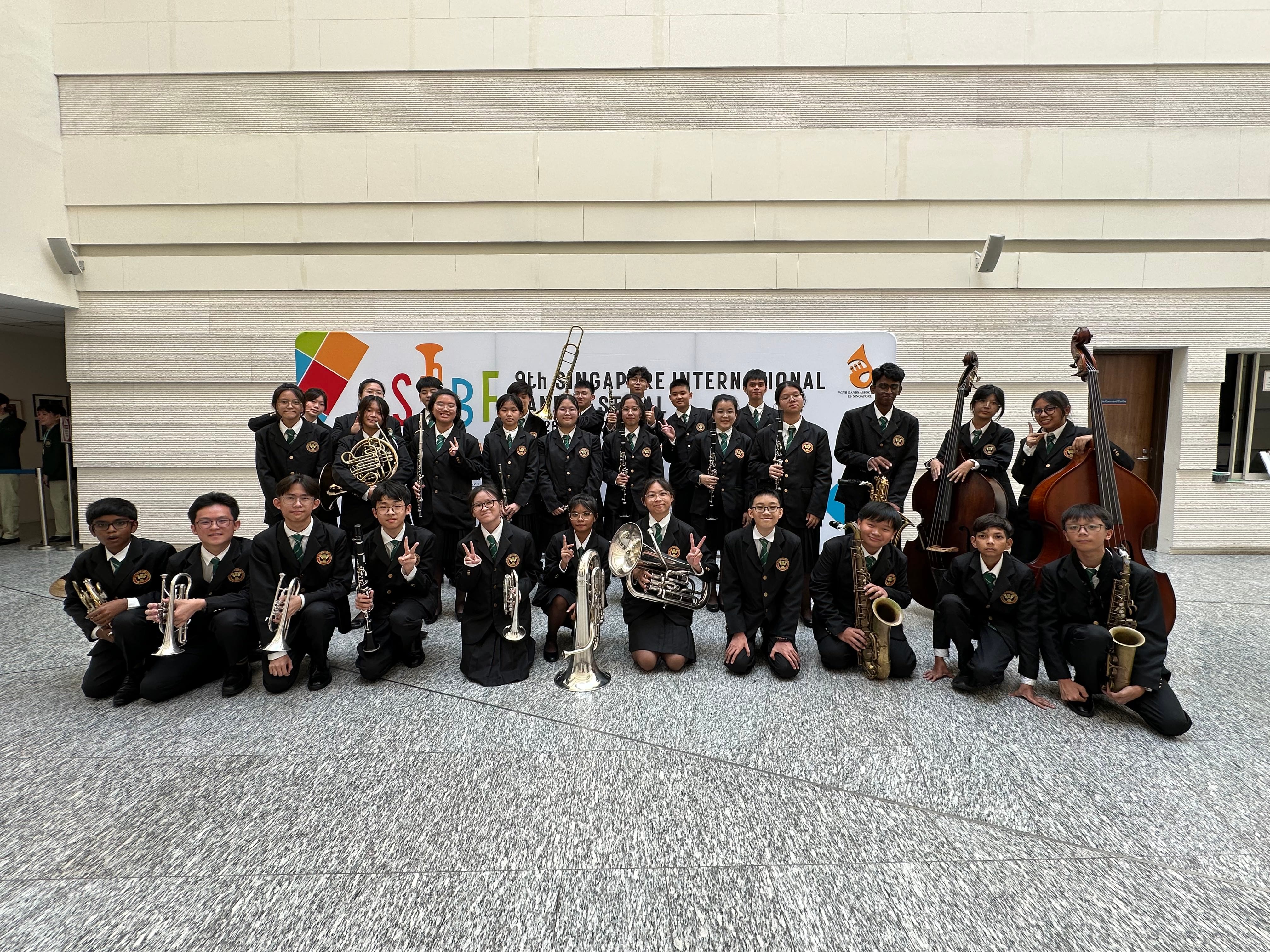 Student band members in black uniforms pose with instruments before a 9th Singapore International Band Festival banner.