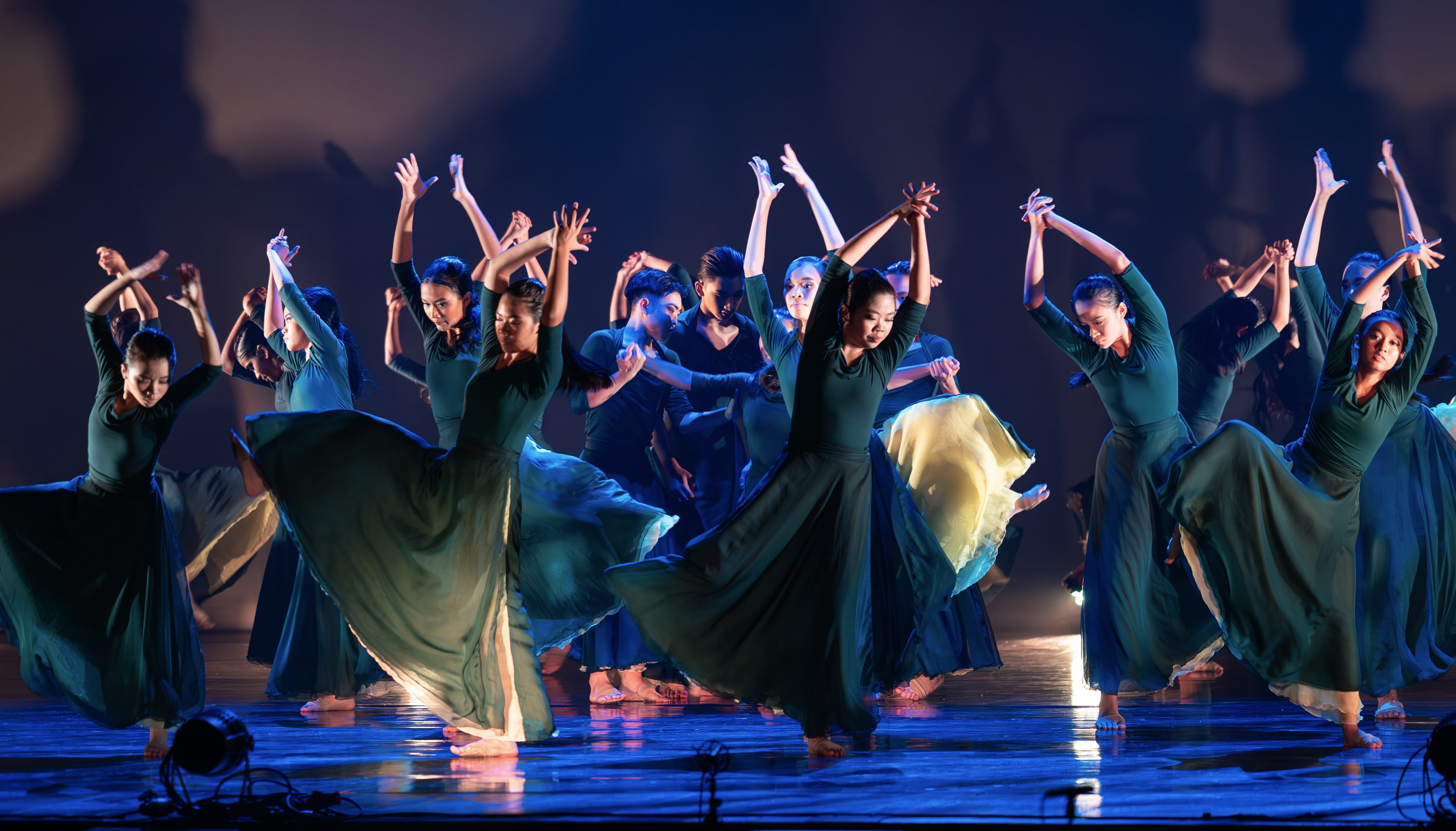 A large group of dancers in flowing teal gowns perform on a dimly lit stage with arms raised.
