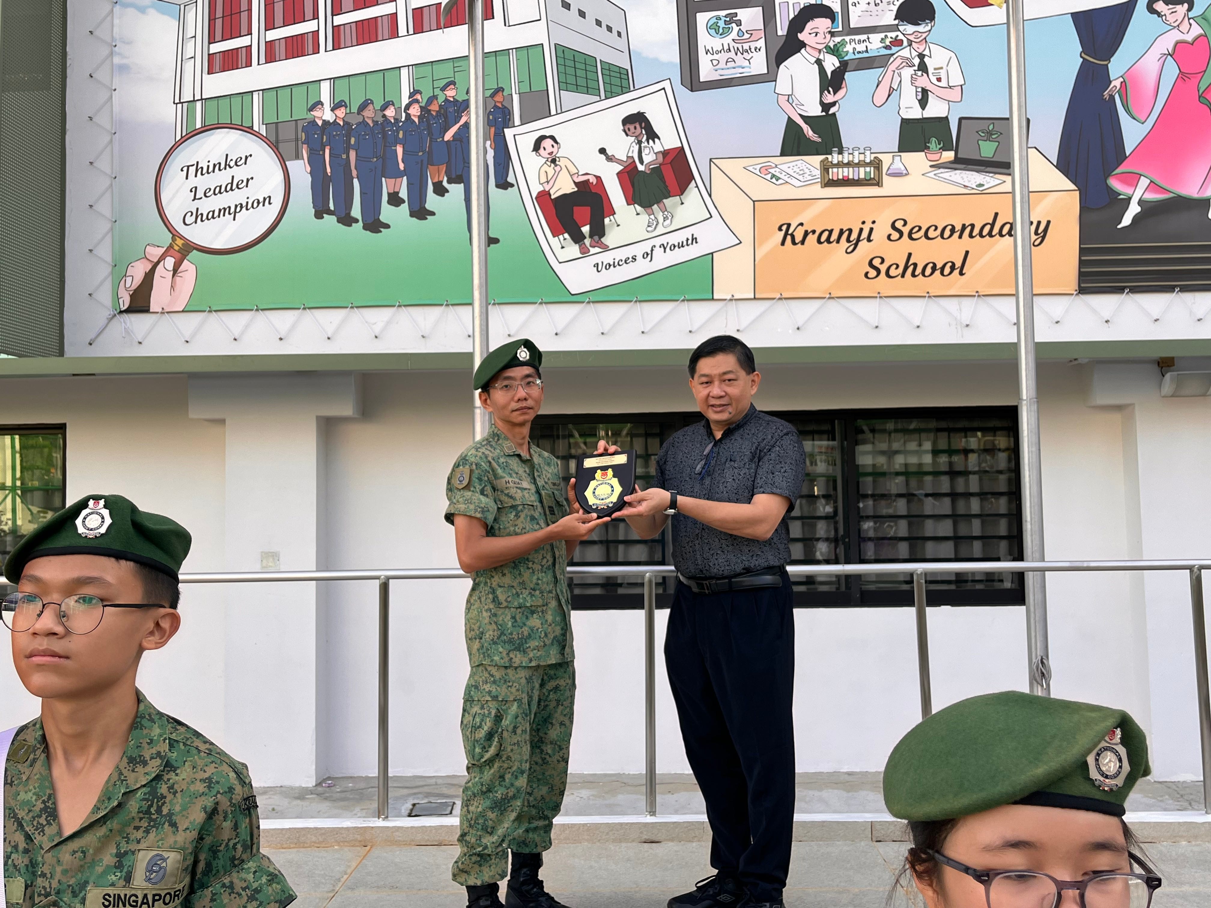 Two men hold a plaque together outside Kranji Secondary School, with uniformed cadets in green berets nearby.
