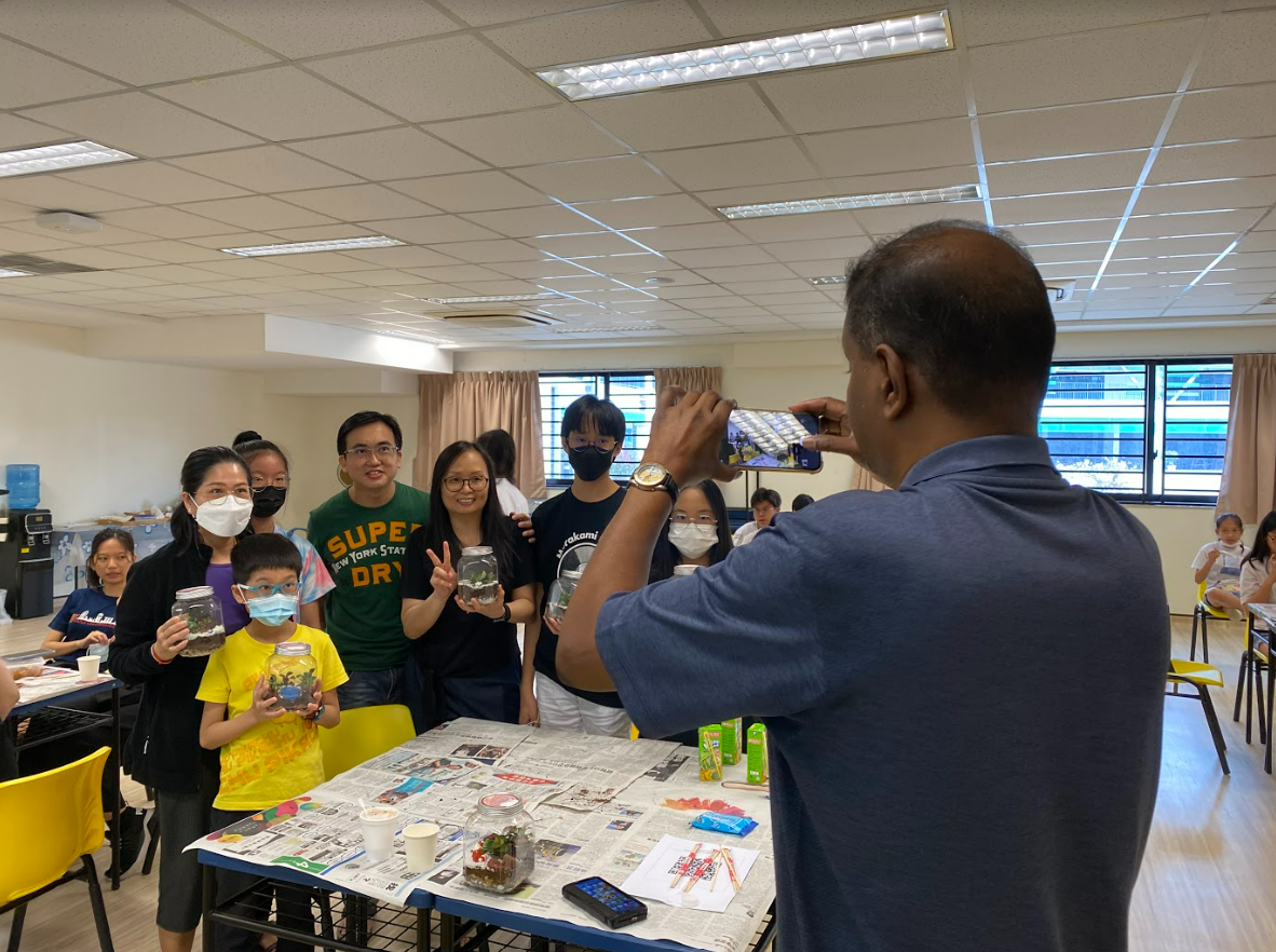 A man photographs a group of people holding glass jar terrariums in an indoor community room.