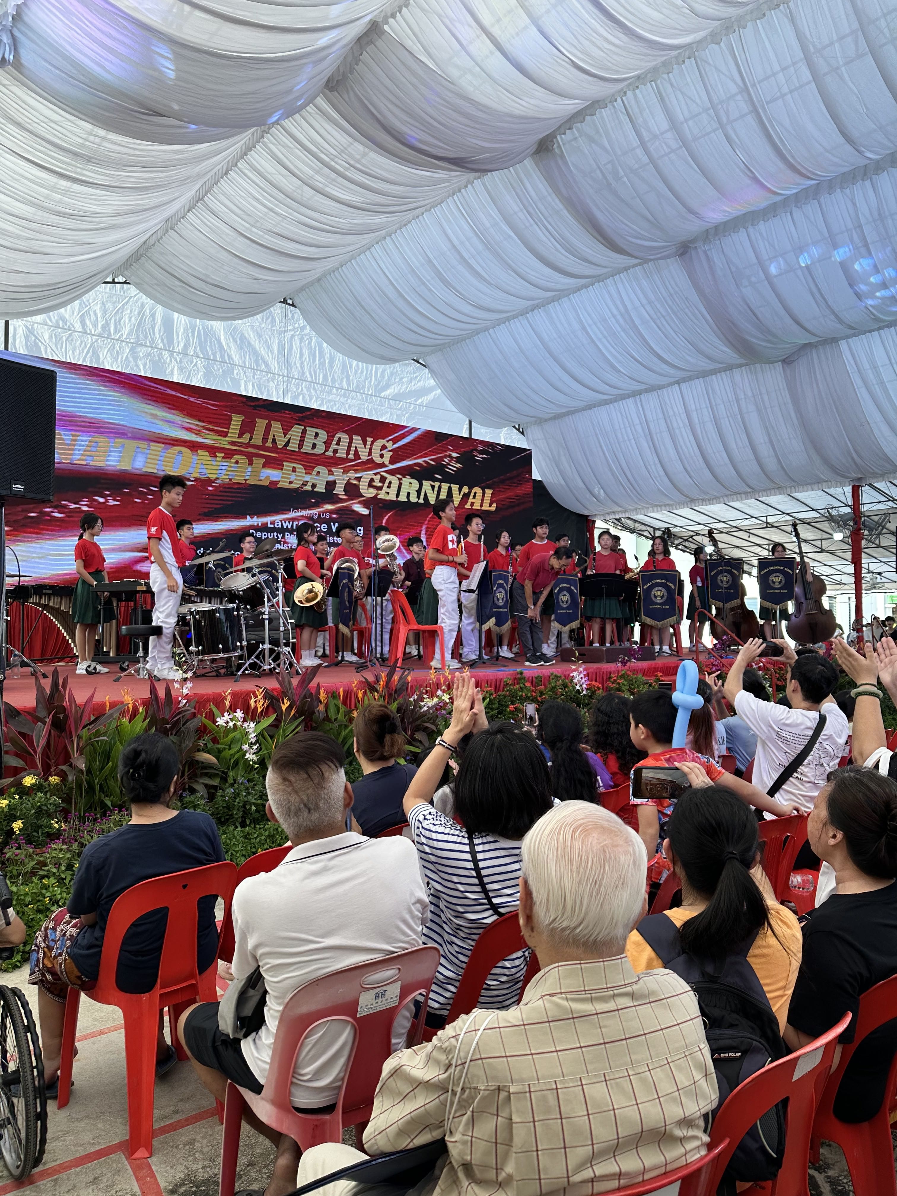 A student band in red shirts performs on stage at the Limbang National Day Carnival as seated audience members applaud.