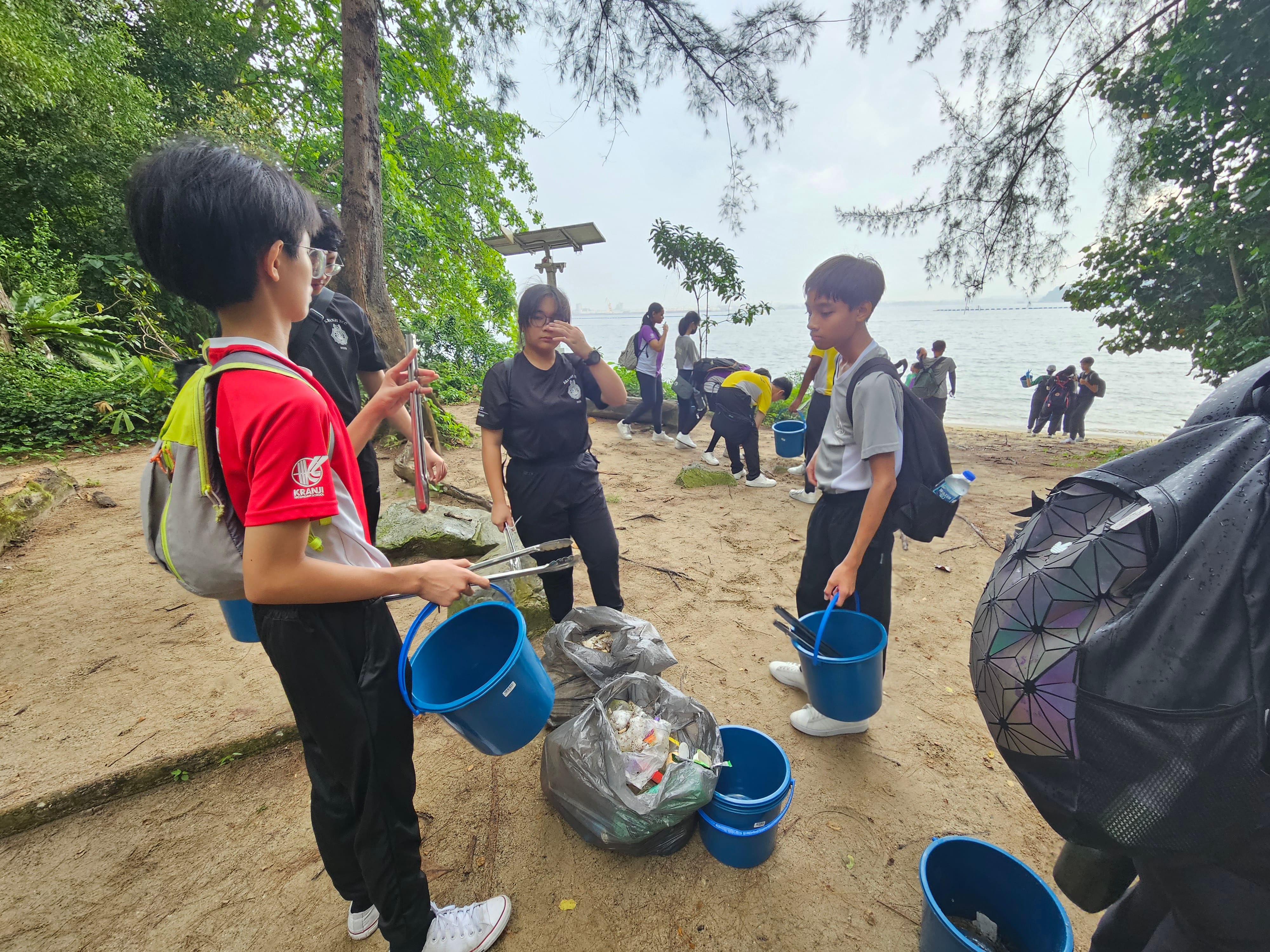 Students on a beach collect litter with grabbers and blue buckets; trash bags piled in the foreground.