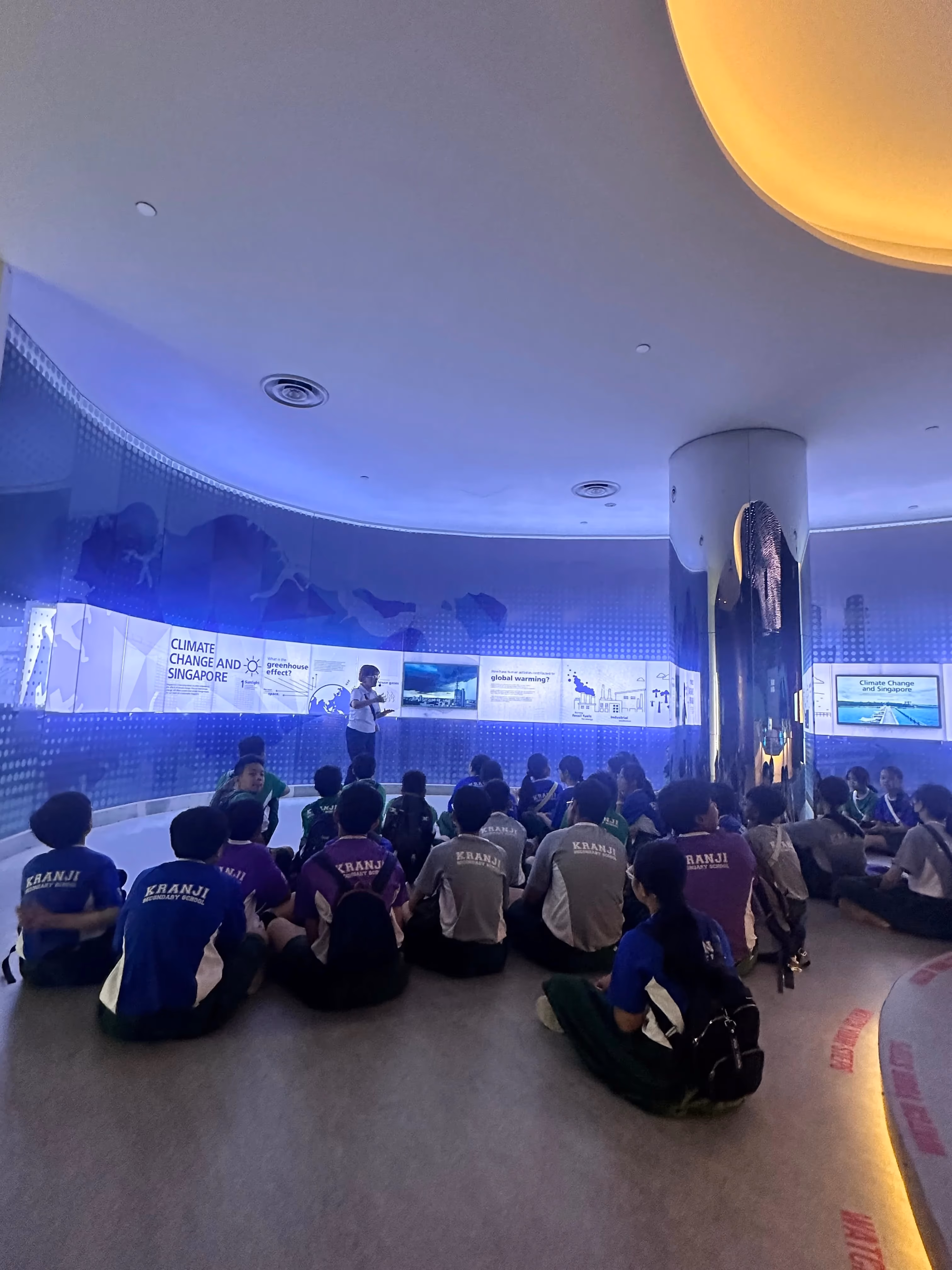 Kranji Secondary School students sit on the floor facing a guide presenting a Climate Change and Singapore exhibit.