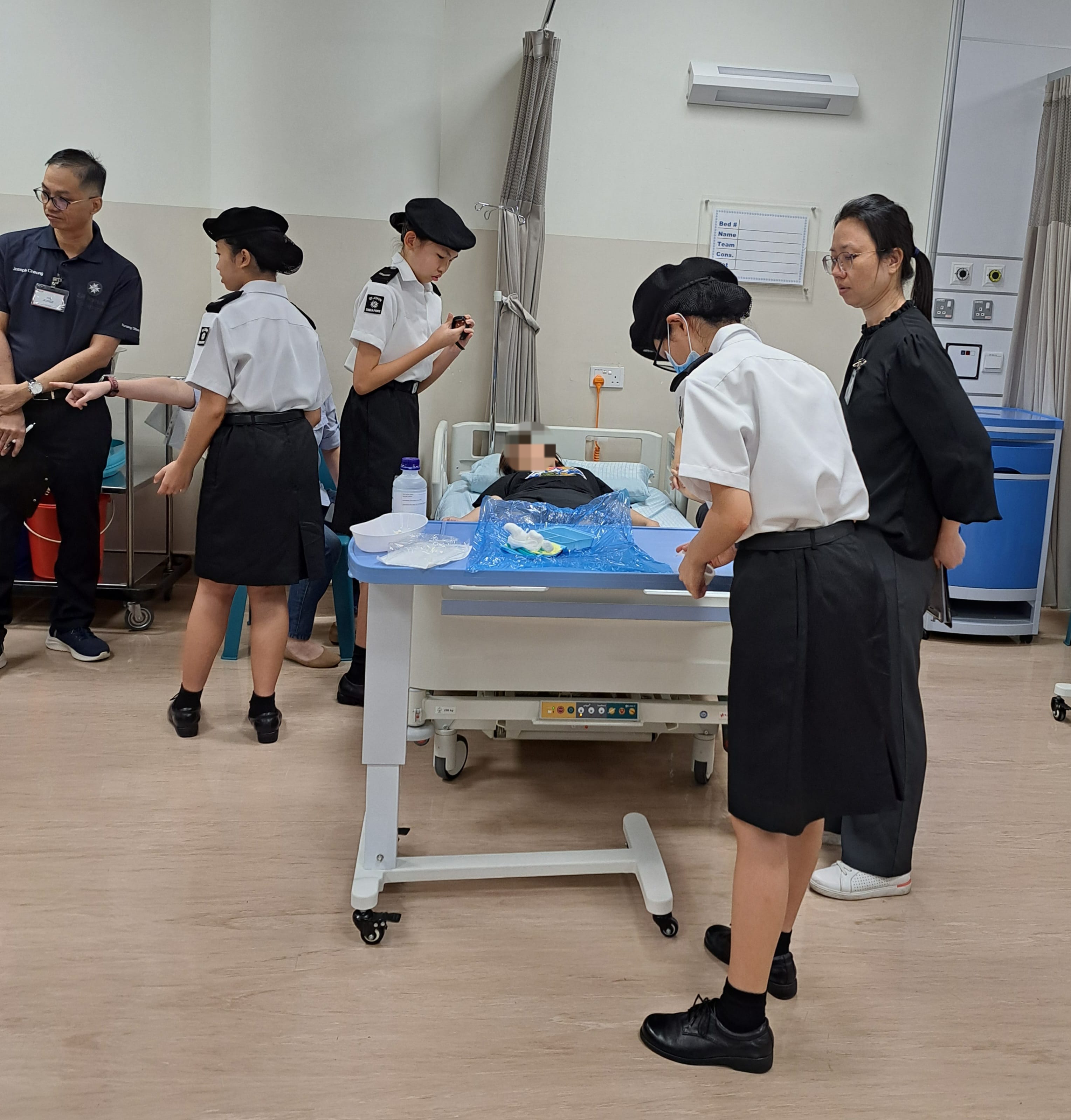 Students in uniform gather around a hospital bed with a patient during a medical training exercise indoors.