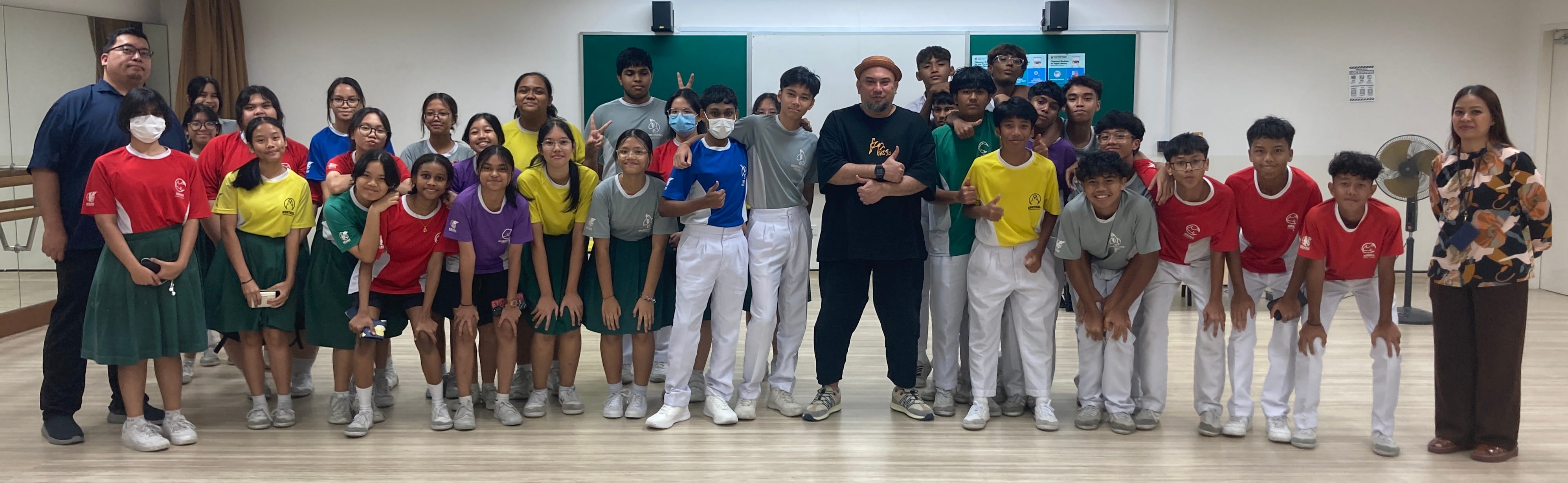 A large group of students in colorful uniforms and two adults pose together in a classroom with a green chalkboard.