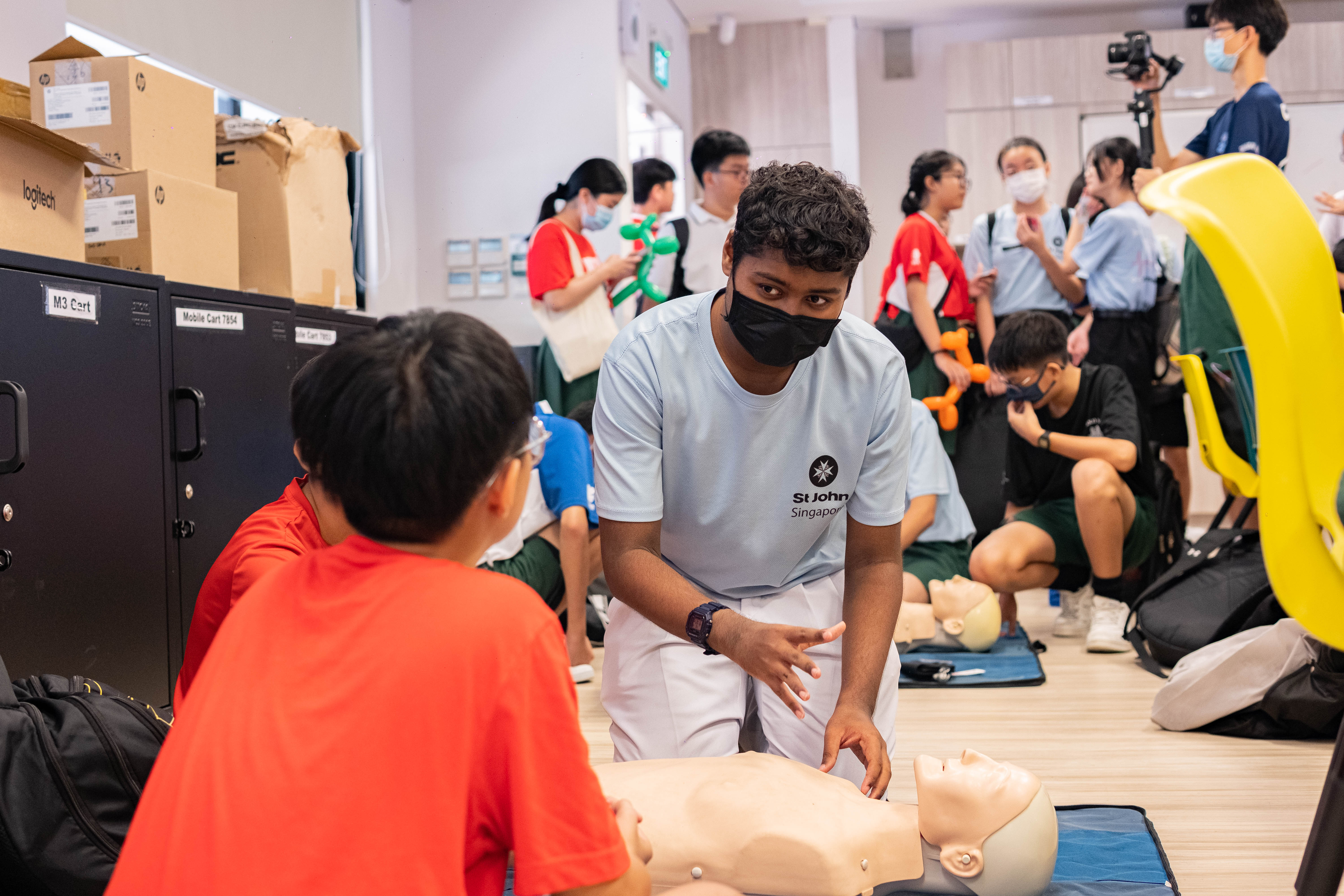 A St John Singapore volunteer in a mask demonstrates CPR on a mannequin to students in a classroom.