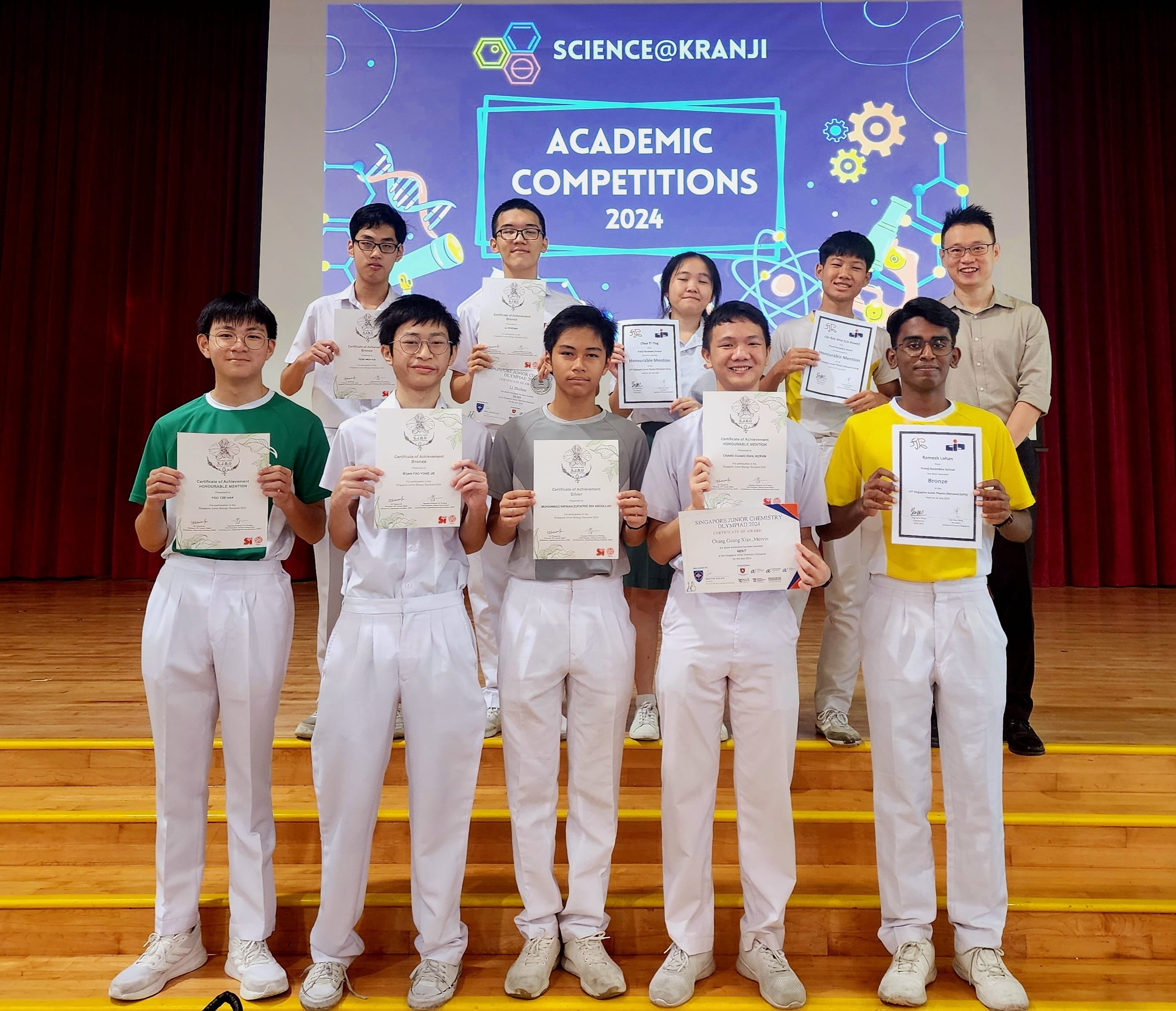 Students hold certificates on a stage in front of a Science@Kranji Academic Competitions 2024 banner.