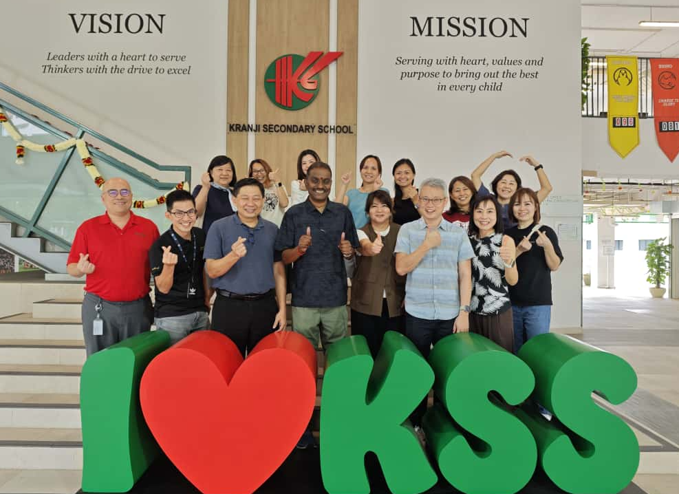 Group of people posing in front of Kranji Secondary School's vision and mission wall with an I Love KSS sign.