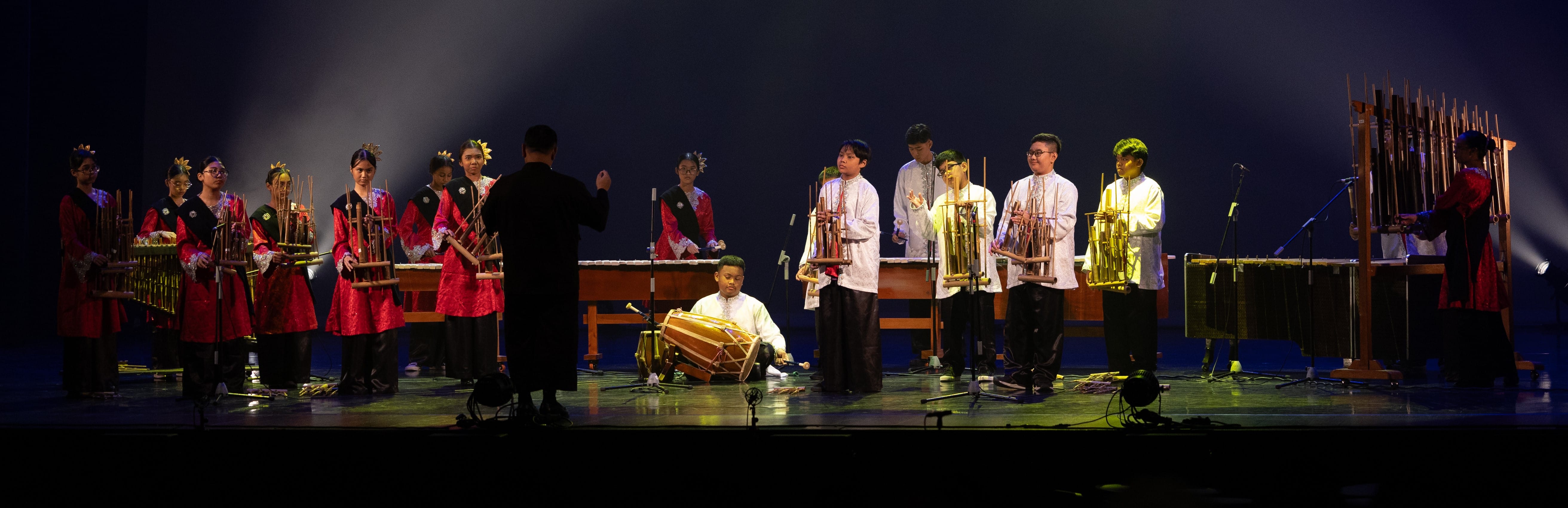 A group of performers in traditional red and white costumes play angklung and percussion instruments on a dimly lit stage.