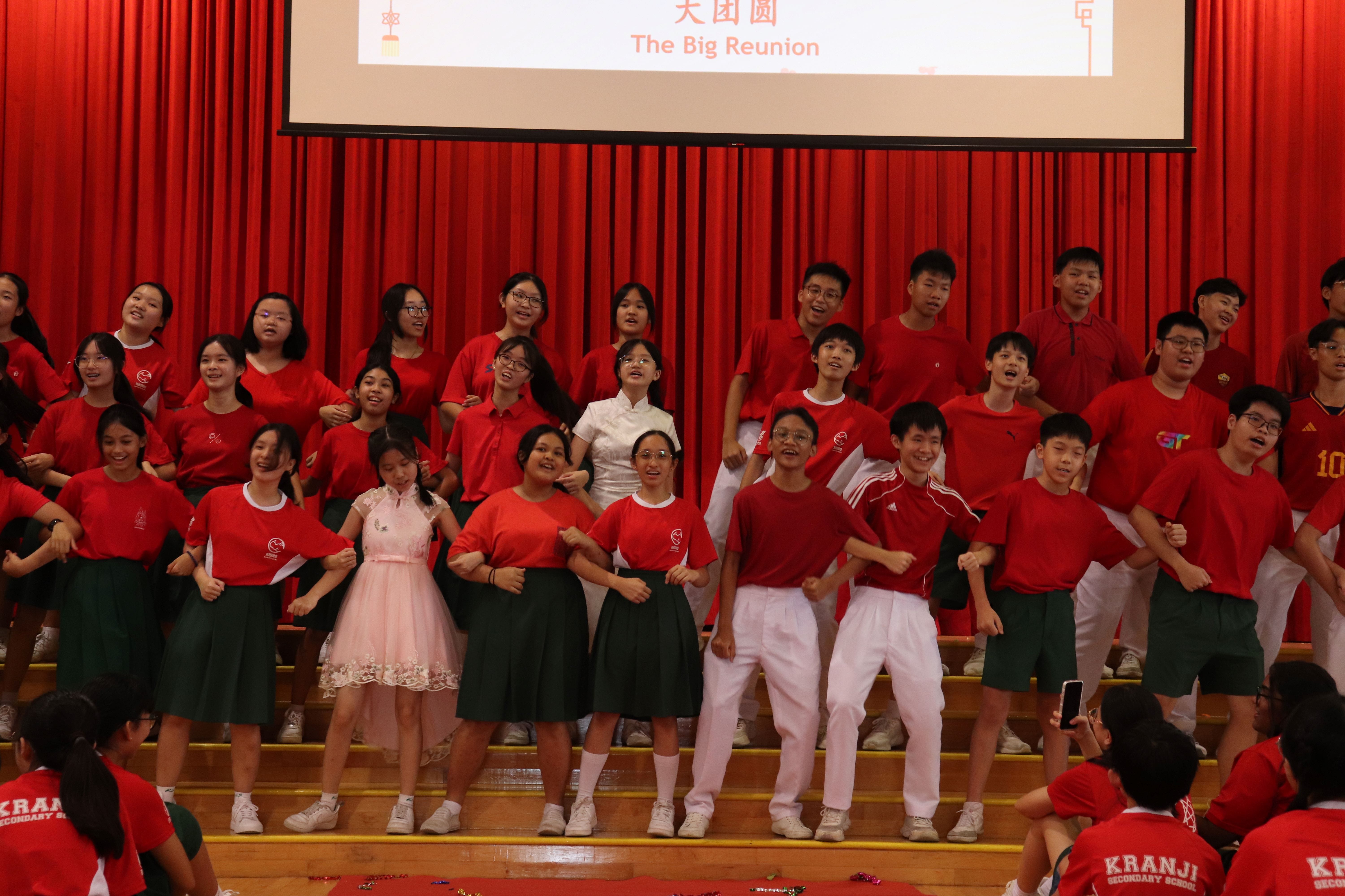 Students in red uniforms perform on a stage with a red curtain and a screen reading "The Big Reunion."