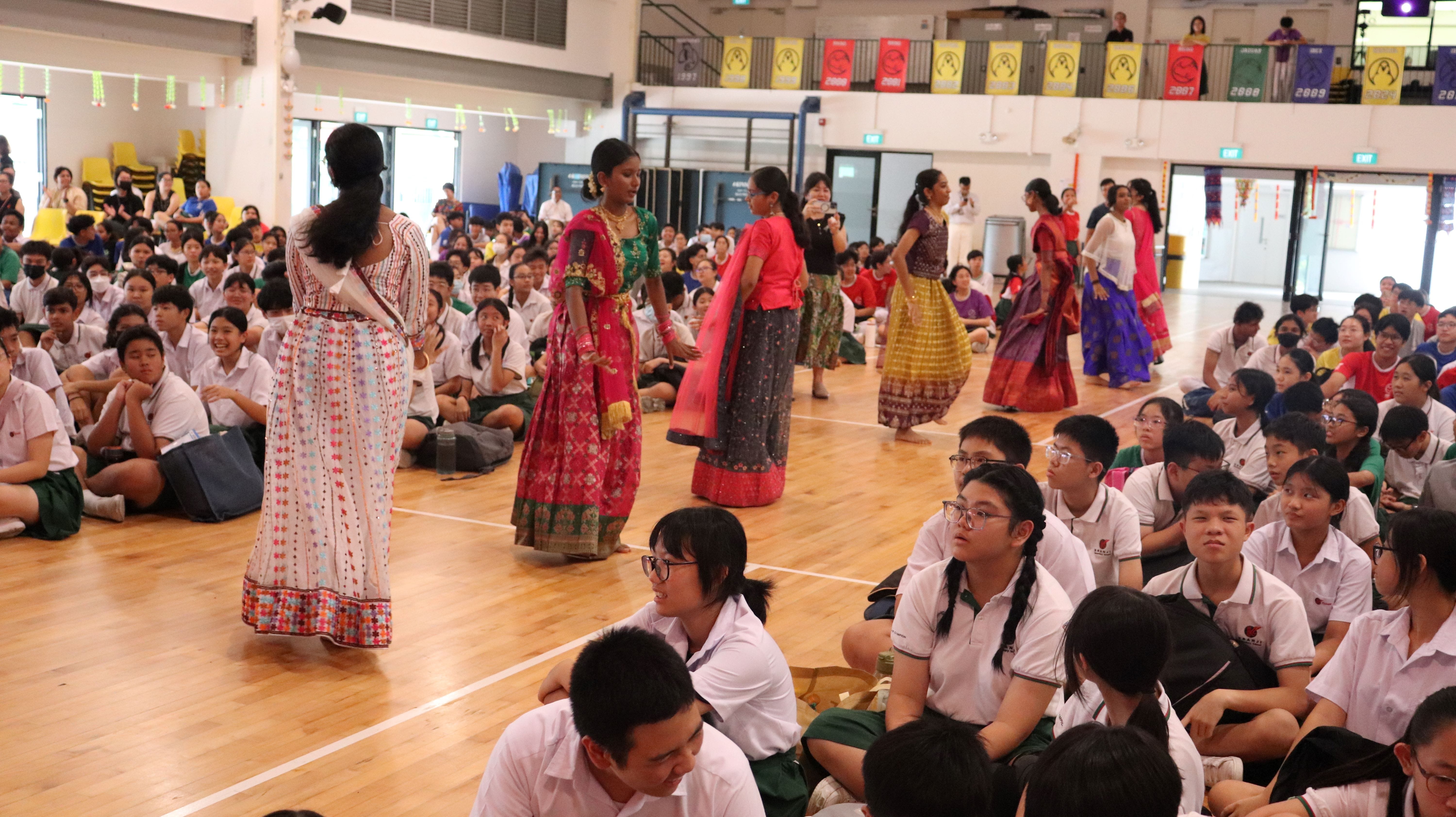 Girls in colorful traditional Indian attire perform in a school hall while uniformed students sit watching on the floor.