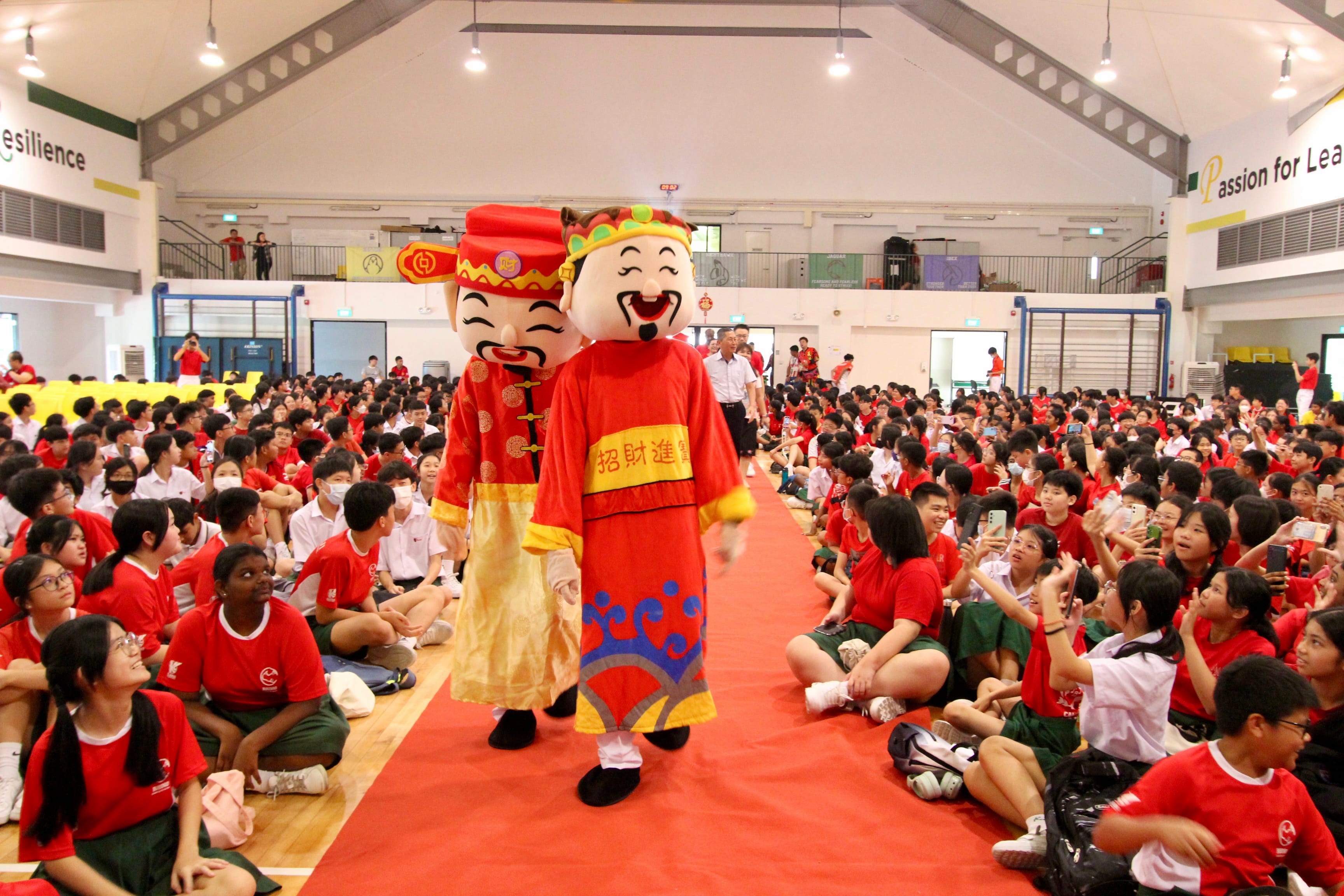 Two Chinese New Year God of Wealth mascots walk a red carpet as students in red shirts sit and watch in a school hall.