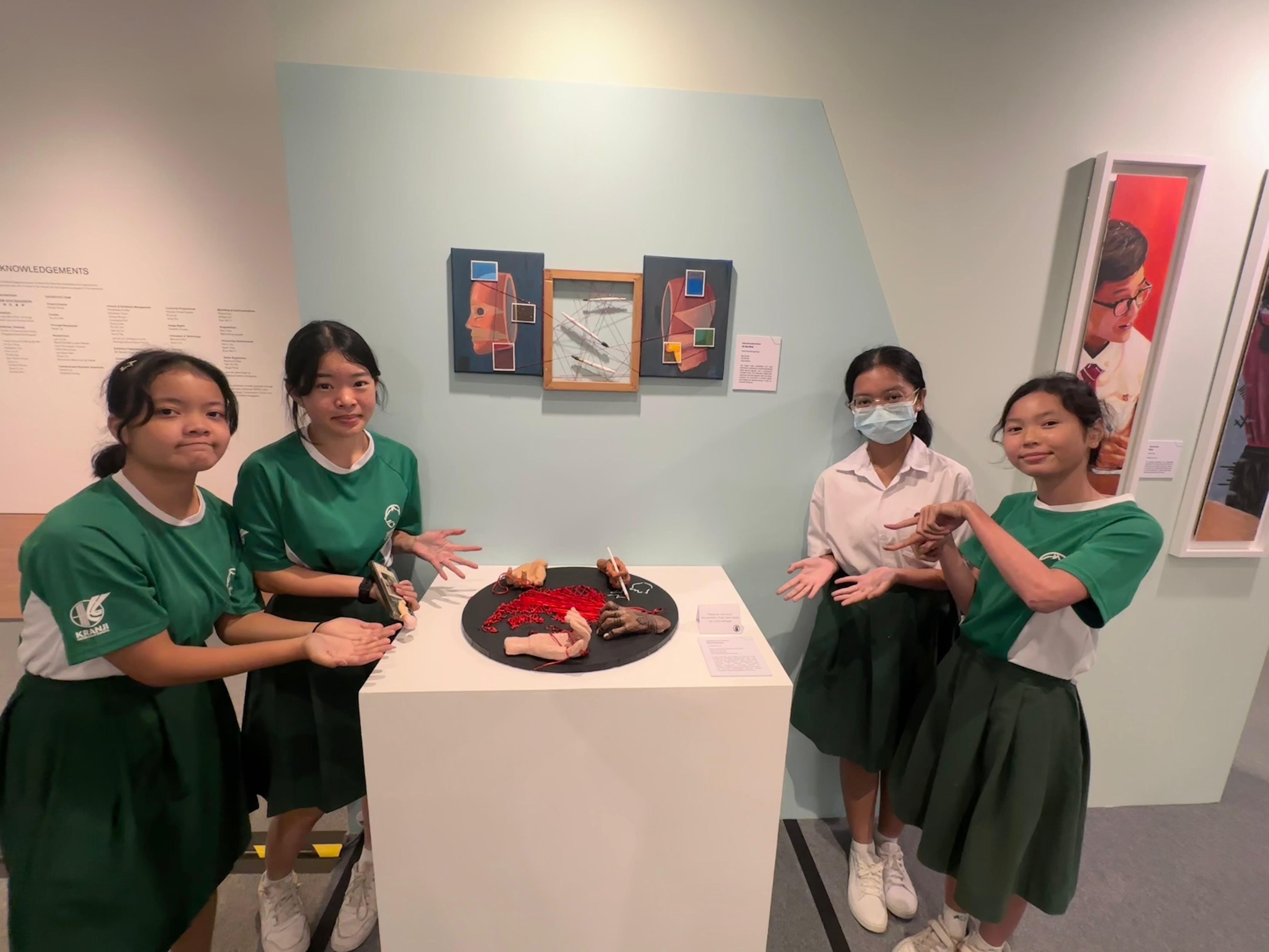 Four school girls in green uniforms pose around a white pedestal displaying a sculpture at an art exhibition.