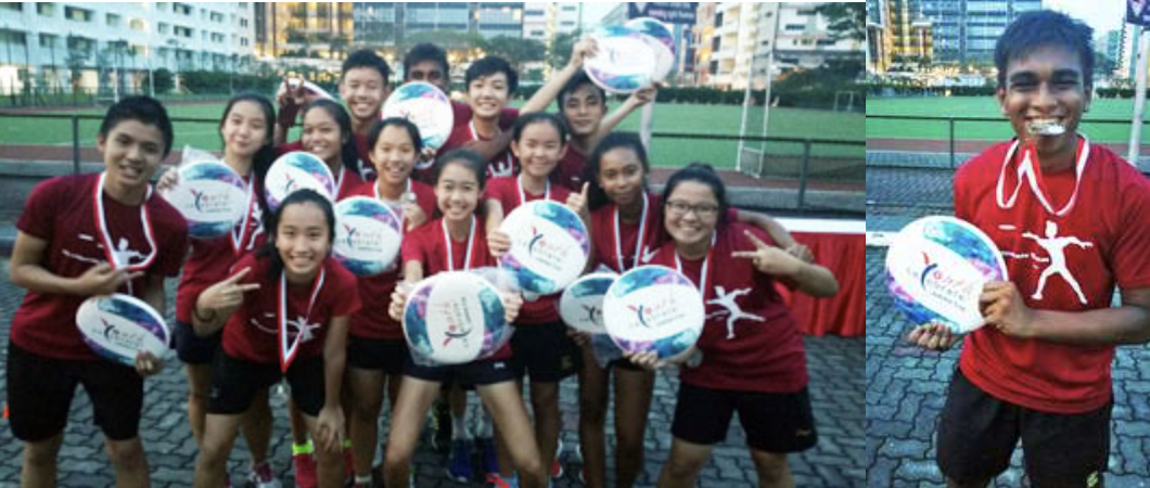 A group of young athletes in red shirts holding round awards and wearing medals, posing outdoors near a sports field.