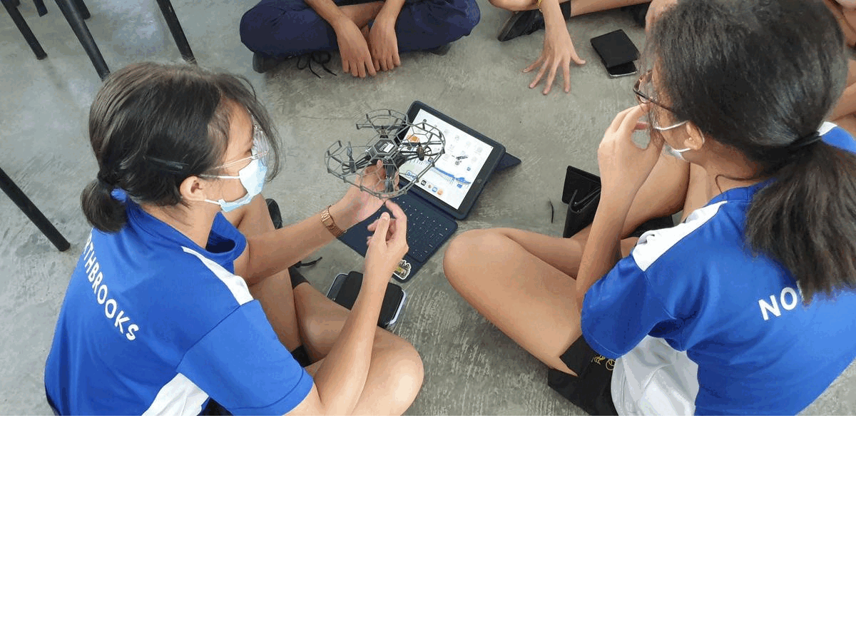 Two masked students in blue Northbrooks shirts sit on the floor handling a small drone and a tablet.