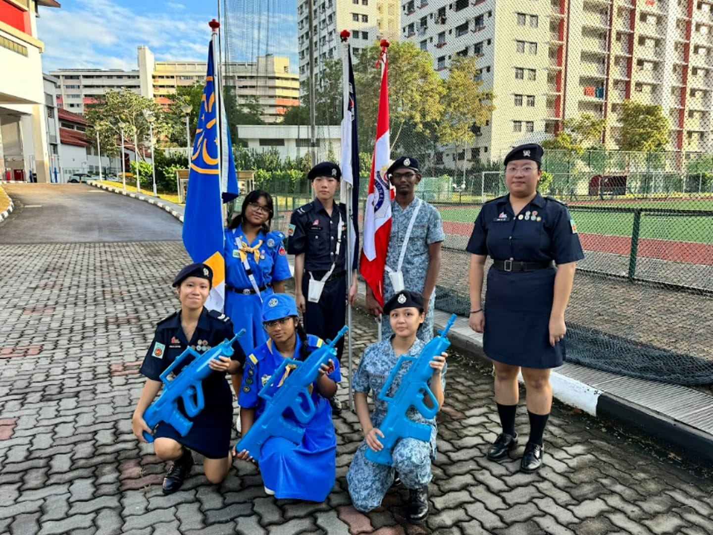 Seven youth cadets in uniforms pose outdoors with flags and blue replica rifles on a paved area near apartment buildings.
