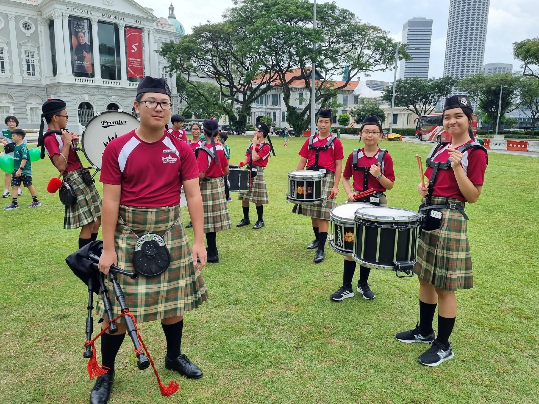 Youth pipe band members in red shirts and tartan kilts holding drums and bagpipes on a grass field near Victoria Memorial Hall.