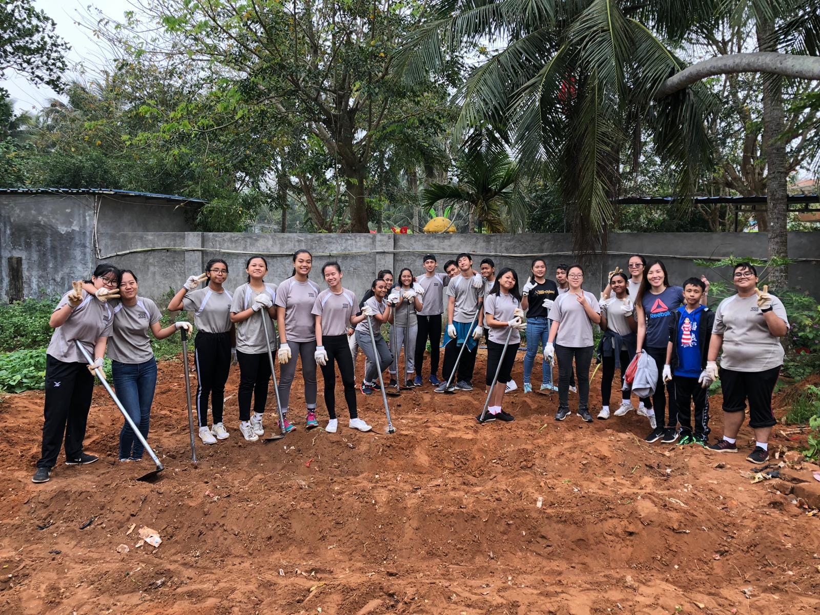 A large group of young volunteers in matching gray shirts pose with shovels on a dirt plot surrounded by trees.