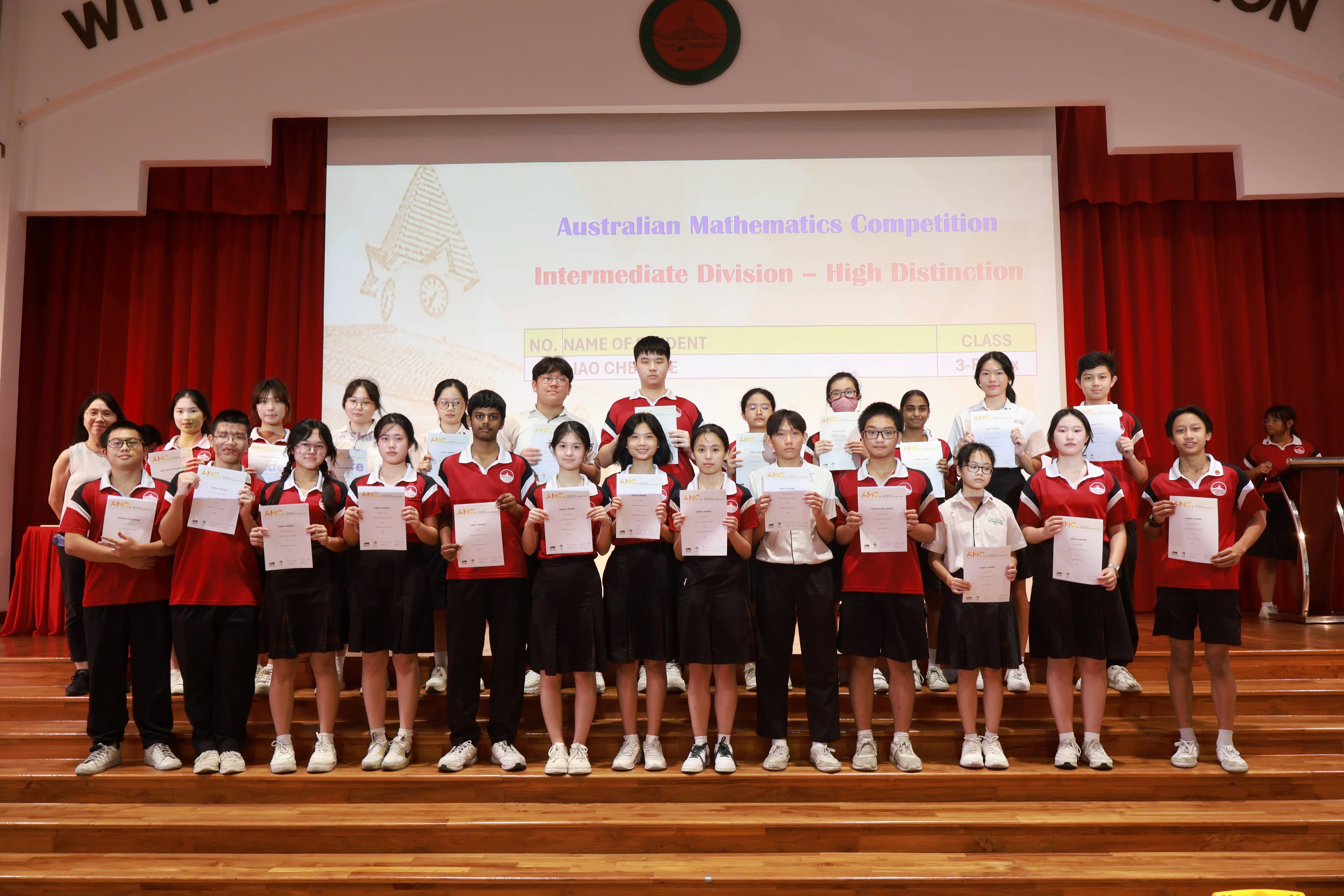Students in red uniforms hold certificates on a stage at an Australian Mathematics Competition High Distinction ceremony.