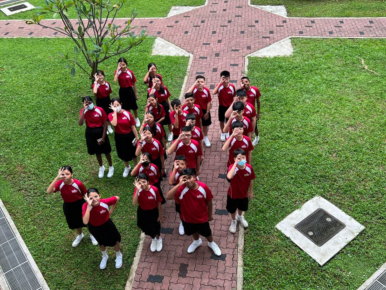 Group of students in matching red and white uniforms posing with OK hand gestures on a brick pathway surrounded by grass.